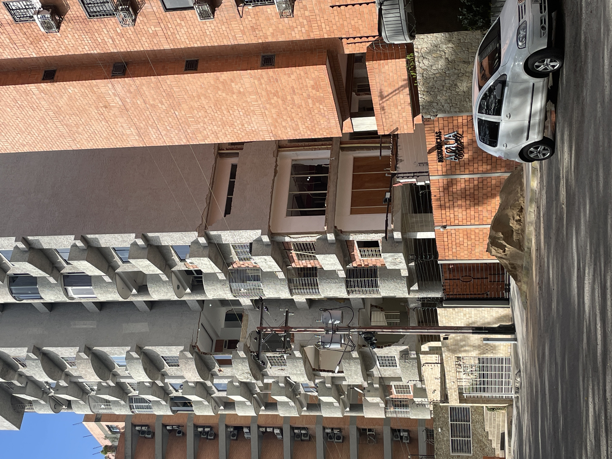 The image depicts a street view in Maracay, Venezuela, focusing on the facades of multiple apartment buildings. The buildings exhibit varied architectural styles and materials, with one prominently featuring grey concrete with rounded balconies and window frames, while another next to it is constructed with orange bricks. A third, older building with a pinkish facade is visible in the background to the left. The scene is predominantly urban, with visible elements of residential infrastructure including windows, air conditioning units mounted on building exteriors, and electrical poles with transformers and wires.
In the foreground, a white Volkswagen Fox car is parked on the paved street. To the right of the car, a low orange brick wall with a sign reading "RESIDENCIAS AQUA" (Residences Aqua) is visible, suggesting the name of the residential complex. Behind the wall, a textured stone facade of a building can be seen. A pile of sand is located to the left of the brick wall and sign. Shadows from the buildings and trees are cast across the street, indicating it is daytime. The sky is clear and blue. The overall impression is one of a typical residential street in a Venezuelan city.