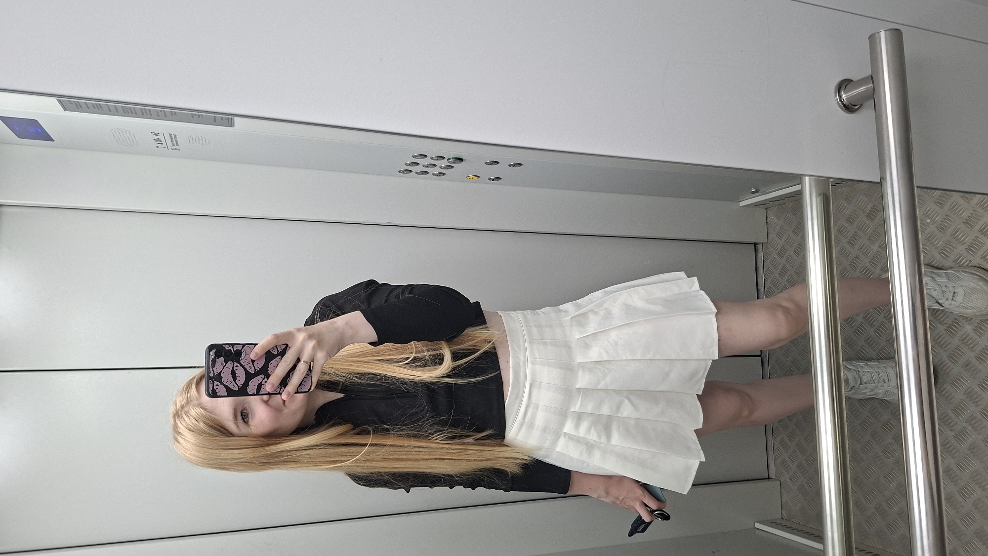 A young woman with long blonde hair takes a mirror selfie in an elevator. She is wearing a black crop top, a white pleated skirt, and white sneakers. She is holding a smartphone with a pink and black lip-patterned case in her right hand. The elevator walls are light gray, and there is a metal handrail in the foreground. The elevator control panel with buttons is visible on the right wall. The image does not contain any information that would indicate the location is in Chaikovsky, Russia.
