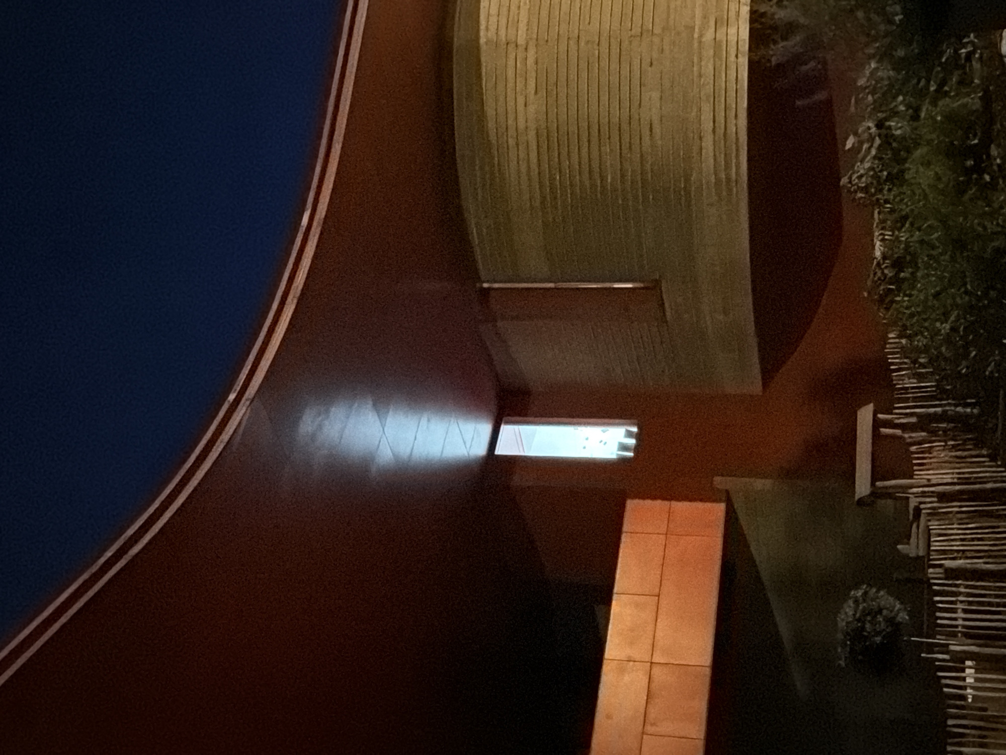 The image depicts an outdoor scene at night, likely in Esch-sur-Alzette, Luxembourg, featuring a section of a curved, reddish-brown structure with a thin white stripe near the top. Below this, a dark blue sky is visible. To the left, a horizontal, orange-toned rectangular element is attached to a darker wall. Further to the right, a curved, light-colored wall with horizontal lines is prominent. In the center, a vertical, narrow window with light emanating from within is set into a dark wall. The foreground consists of dark greenery, a small tree, a rustic fence made of vertical wooden stakes, and what appears to be a small wooden wishing well structure. The overall lighting suggests artificial illumination from within the building and possibly from nearby sources.