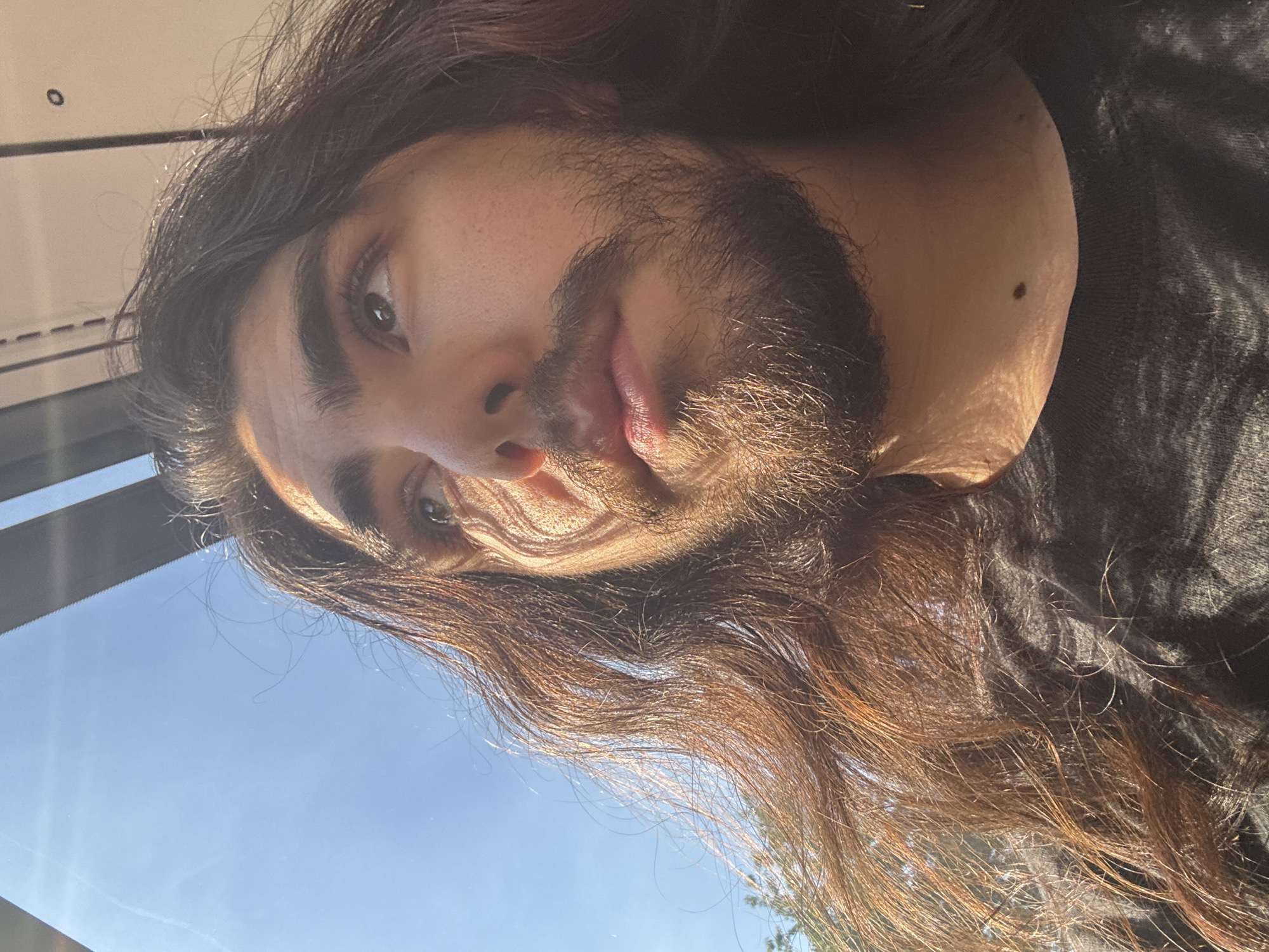 A close-up, low-angle, outdoor, medium shot captures the head and shoulders of a person with long, dark, wavy hair and a beard. The person is wearing a dark t-shirt. The sun illuminates the person from the left, casting shadows across their face. The background consists of a bright blue sky with hints of a wire and a dark, angled structure, possibly part of a vehicle or building. The overall lighting suggests a sunny day.