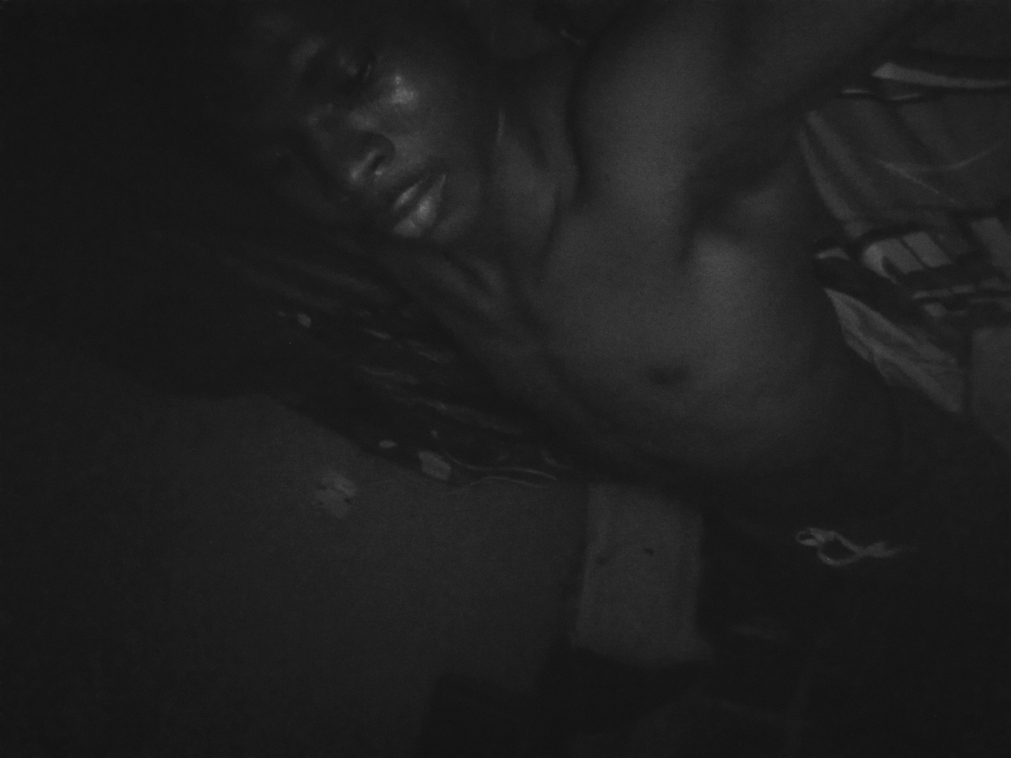 A dimly lit, grainy, black and white image depicts a shirtless person lying down. The subject appears to be dark-skinned. The person's face is visible. The background is indistinct. The location of Nkpor, Nigeria is not determinable from the visible content of this image.
