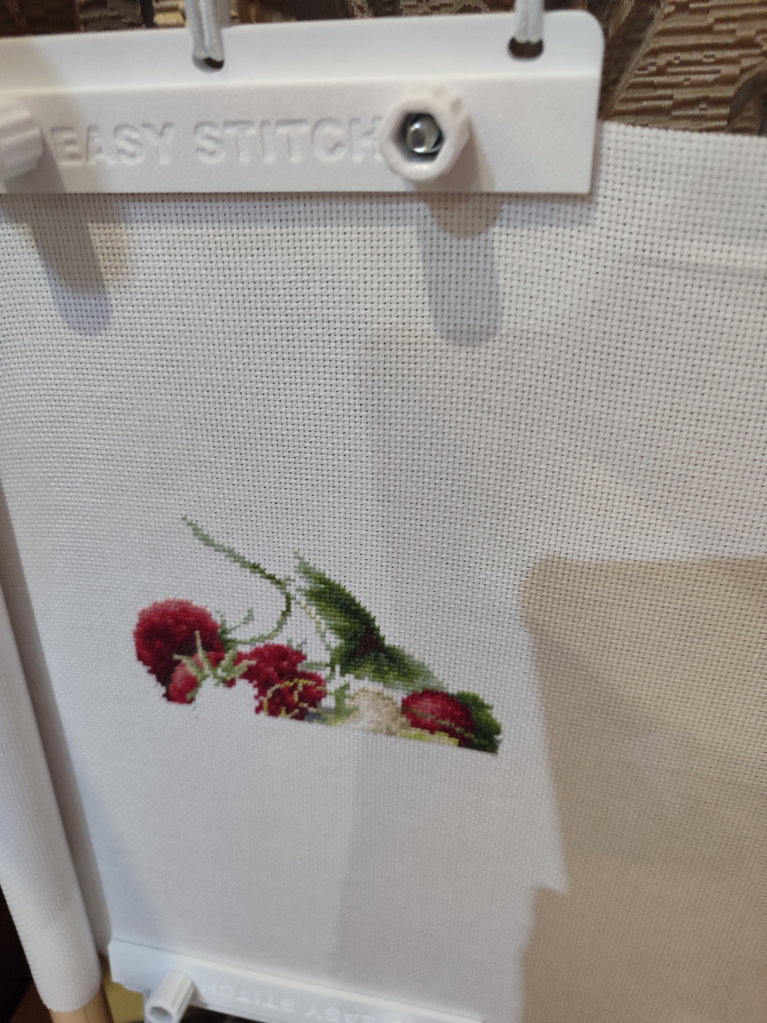 The image shows a close-up of a white cross-stitch fabric stretched on a white plastic frame. The frame has the embossed text "EASY STITCH". The fabric is partially embroidered with red flowers and green leaves. The embroidery is positioned in the lower left quadrant of the frame. The background behind the frame appears to be a textured fabric, possibly upholstery. The lighting suggests an indoor setting. There are no people or any indication of the city of Tuapse, Russia.