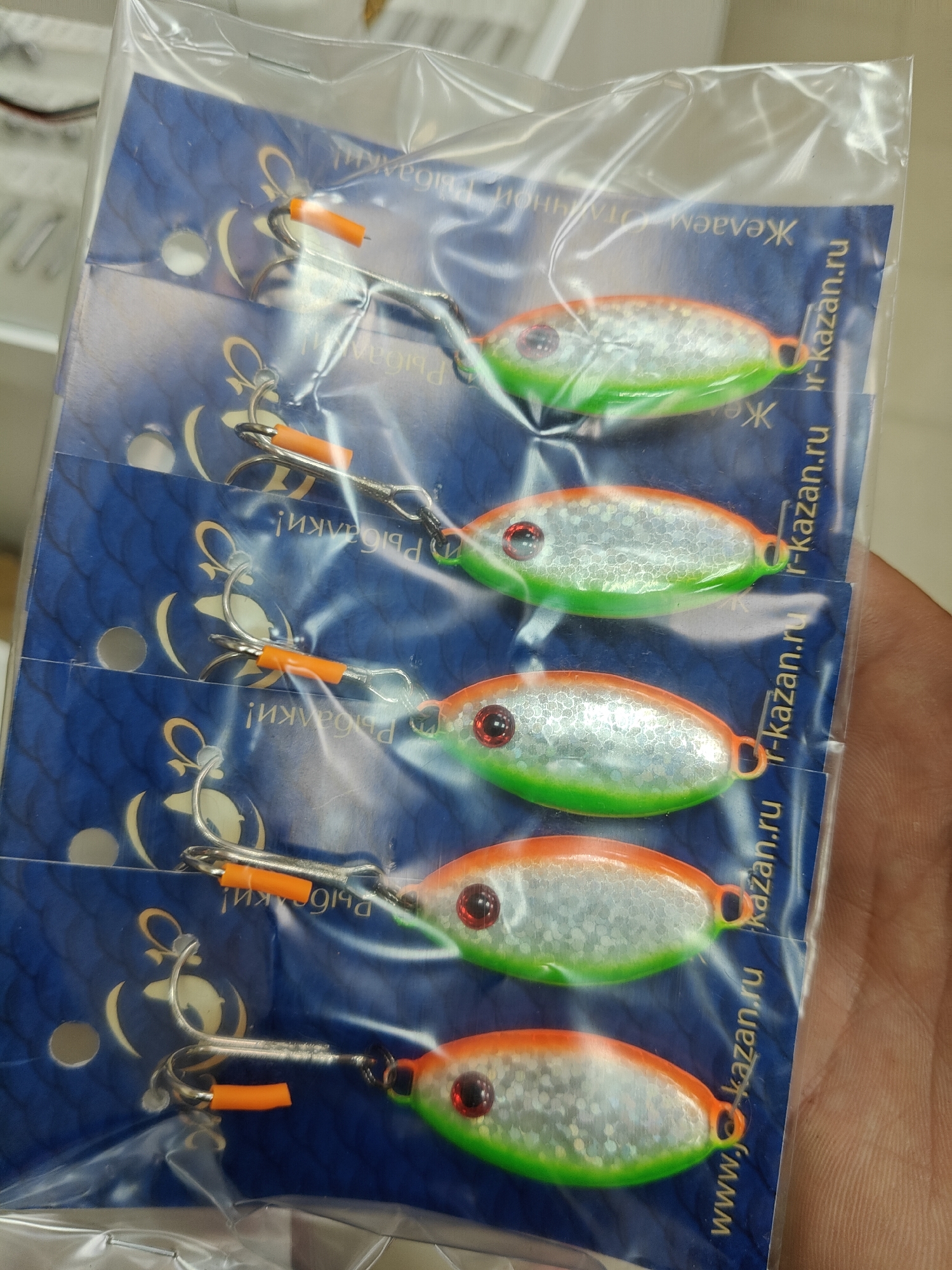 The image shows a package of five fishing lures, each attached to a blue card with Russian text. The lures are teardrop-shaped, with silver bodies, colored edges (green or orange), red eyes, and orange pieces on their hooks. The cards are stacked within a clear plastic package, which is being held by a hand. The website address "www.j-kazan.ru" is printed on the blue cards. The text on the card, "Рыбалки!", translates to "Fishing!". The image was taken in the city of Chaikovsky, Russia.