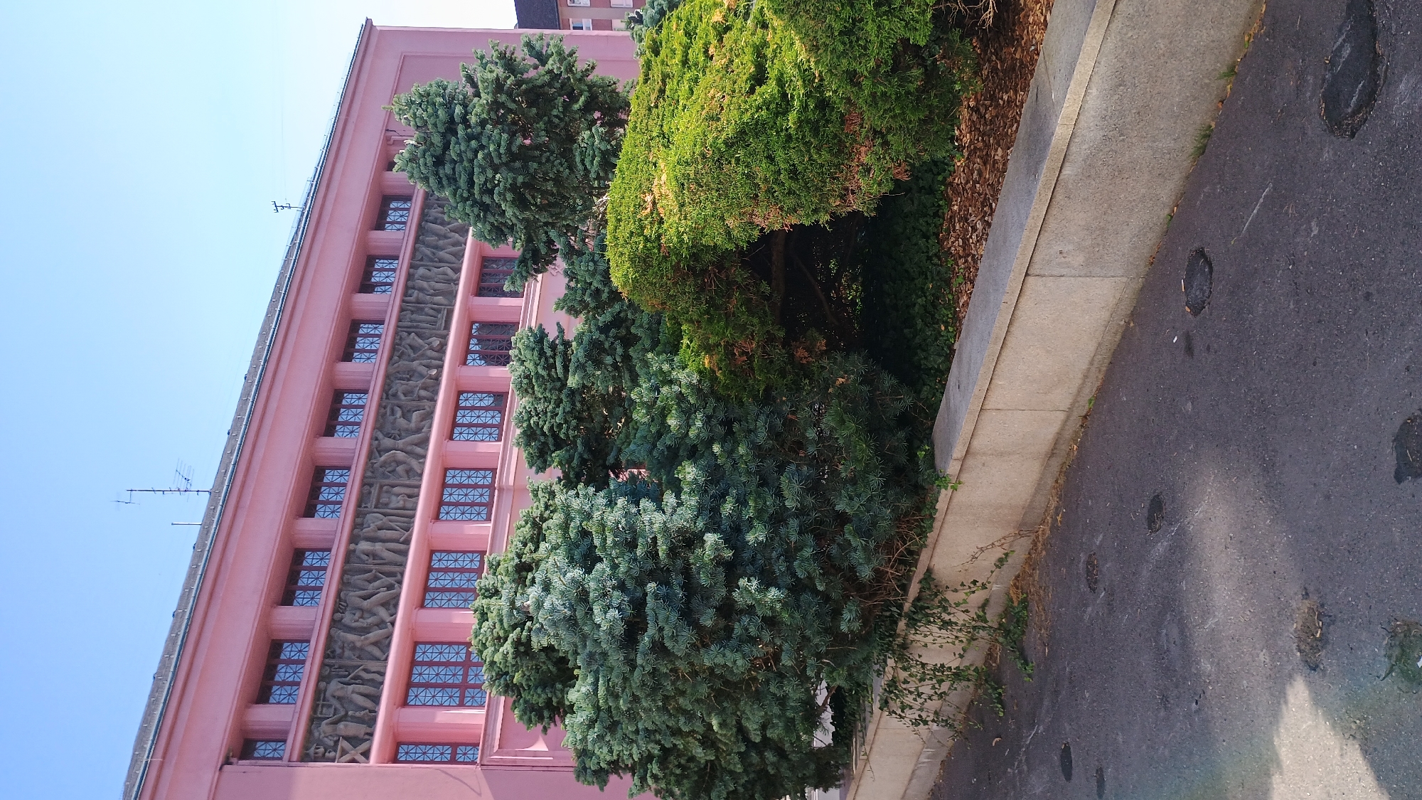 The image captures a facade of a pink building with a frieze of relief sculptures depicting human figures in an industrial setting. The building features rows of windows with intricate lattice patterns. In the foreground, lush green bushes and trees create a natural barrier. A concrete wall separates the vegetation from an asphalt road, which shows several circular manhole covers and patches of wear. The sky is clear and blue, indicating a sunny day. The architectural style and the relief sculptures suggest a period of socialist realism, often found in public buildings constructed during the communist era in Eastern Europe. While the specific location is not visually evident, such architecture is characteristic of many cities in the Czech Republic that underwent significant development during the mid-20th century.