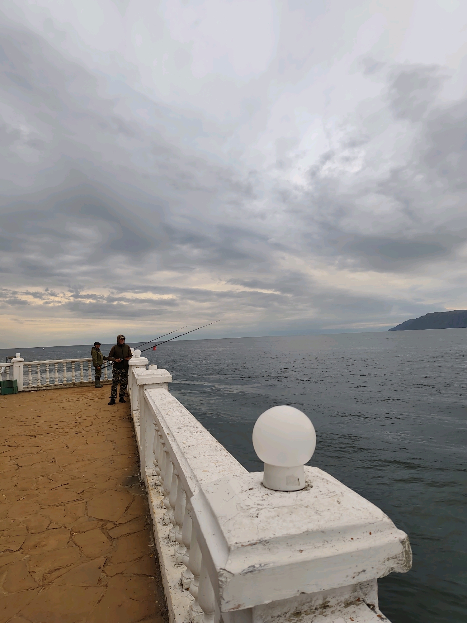 The image depicts an outdoor scene on a pier in Listvyanka, Russia, a settlement on the southwestern shore of Lake Baikal. The sky is overcast with a gradient of gray clouds. The sea is a dark, choppy blue. In the foreground, a white balustrade with ornate spindles runs along the edge of the pier. A spherical white lamp is mounted on the balustrade in the lower right corner. The pier itself is paved with irregular, flat, light brown stones. Further down the pier, two men are fishing. The man on the right is wearing a green hooded jacket and camouflage pants and is holding a fishing rod. The man on the left is wearing a green jacket and light-colored pants and is also holding a fishing rod. Their fishing rods are extended out over the water. A white decorative railing with balusters borders the pier on the left side, separating it from the water. In the distance, a hazy, dark green landmass, likely a mountain range, is visible along the horizon. A small white object, possibly a boat, is visible on the water to the far left. The overall impression is one of a cloudy, possibly cool day at the water's edge.