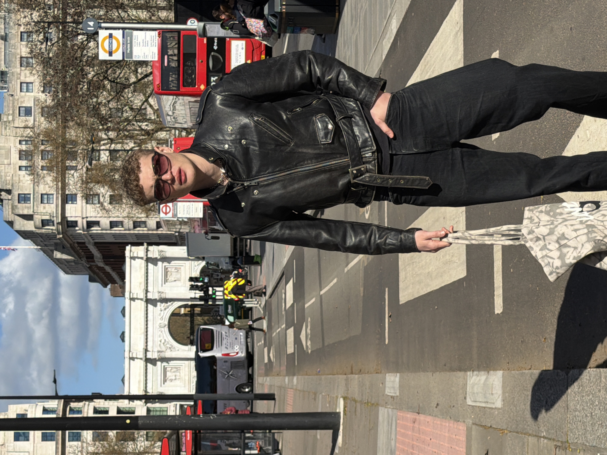 The image captures an urban street scene in London, United Kingdom, featuring Marble Arch. A young male subject stands in the immediate foreground, positioned on a pedestrian crossing, looking directly towards the viewer. He wears a black leather biker jacket with a belted waist, dark oval sunglasses, a dark shirt, and black jeans. His left hand holds a light-colored, patterned tote bag, while his right hand rests in his front jeans pocket. Strong shadows cast by the subject and other objects indicate bright daylight conditions.
In the midground, the grand white neoclassical Marble Arch is prominently visible. Surrounding the arch and along the street are multi-story stone buildings. Traffic on the road includes a red double-decker bus partially visible on the right, a silver coach bus driving away to the left, and a white truck. Several pedestrians are present on the sidewalks and crossing the road, including individuals in a green jacket and a red top. Bare trees with no foliage are visible between buildings. A bus stop sign identifying "COACHES," "Marble Arch," and route "14" is located on the right side. The sky is blue with scattered white clouds.