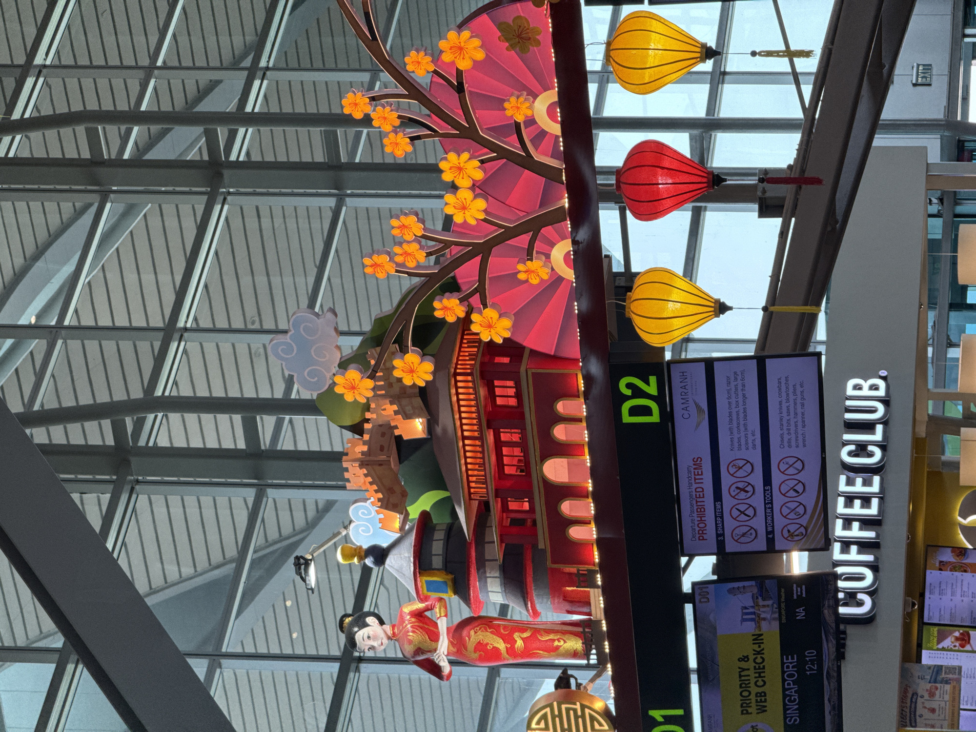 The image shows a close-up, low-angle view of a decorative display with traditional Asian elements, likely at Cam Ranh International Airport in Cam Ranh City, Vietnam. The display features a large, stylized figure of a woman in a red and gold traditional dress, standing next to miniature replicas of traditional Chinese architecture, including a pagoda and a section of the Great Wall. The structures are lit from within and adorned with small orange flowers and decorative clouds. A large, pink fan-shaped backdrop is also visible.
Hanging below the display are three traditional Asian lanterns: two yellow and one red, each with decorative tassels.
In the background, elements of the airport's interior are visible, including large windows, metal structural beams, and signage. Two digital display screens are prominent. One shows "PRIORITY & WEB CHECK-IN" for Singapore, with the time "12:10". The other screen displays "CAMRANH INTERNATIONAL TERMINAL" and lists "PROHIBITED ITEMS" for departure passengers, with categories like "SHARP ITEMS" and "WORKER'S TOOLS," accompanied by cautionary icons. Below these screens, a prominent illuminated sign reads "COFFEE CLUB."
