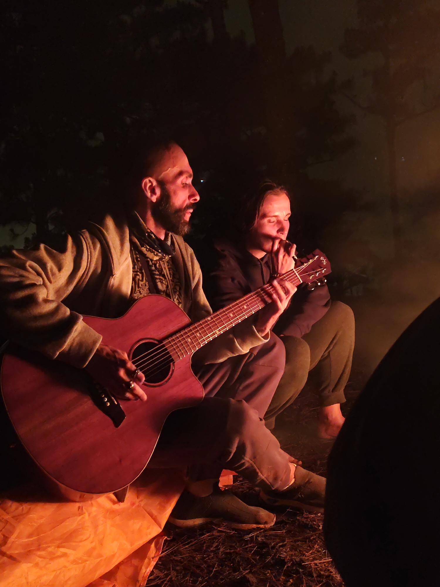 In a nocturnal outdoor scene, two individuals are gathered around what appears to be a campfire, illuminated by its warm, orange glow. The focal point is a man, positioned on the left, playing an acoustic guitar. He has a beard and is wearing a light-colored hooded jacket over a patterned scarf or shirt. Rings are visible on his right hand as he strums the guitar. His expression suggests focus and enjoyment.
To his right, a second person, possibly female, is seated, engrossed in the music. This individual is wearing a dark hoodie and dark pants, with hands clasped near their face, potentially singing or simply listening intently. The guitar has a reddish-brown wooden finish.
The ground around them is covered with dry foliage, possibly leaves or pine needles, suggesting a natural, forested environment. An orange tarp or cloth is spread on the ground near the guitarist, reflecting the firelight. The background is largely dark, with hints of tree silhouettes against the night sky, further emphasizing the isolated and intimate setting. The light source, presumably a campfire, casts strong shadows and highlights, creating a cozy and atmospheric mood. No text is visible in the image. This serene gathering takes place in Lạc Dương, Vietnam.
