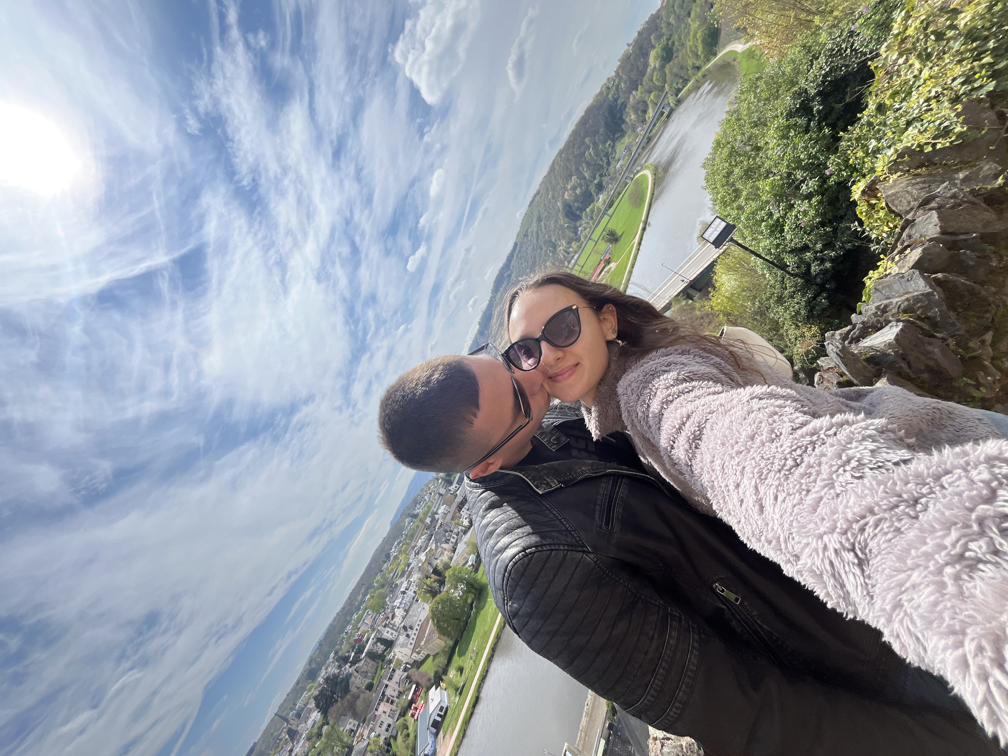 A couple takes a selfie overlooking Saarburg, Germany, on a bright, sunny day with a blue sky dotted with clouds. The perspective is canted to the left, placing the sky and cityscape at an angle.
The woman, positioned closer to the camera, wears sunglasses and a soft, light gray jacket. The man, slightly behind her, is in a black leather jacket and eyeglasses. They are close together, sharing a kiss or near-kiss pose.
In the background, Saarburg is visible with its clustered buildings and the Saar River winding through the town. Green hills and vegetation frame the river. A bridge crosses the river. In the immediate foreground, there are rocks and greenery. The sun is positioned in the upper left, causing a bright glare.