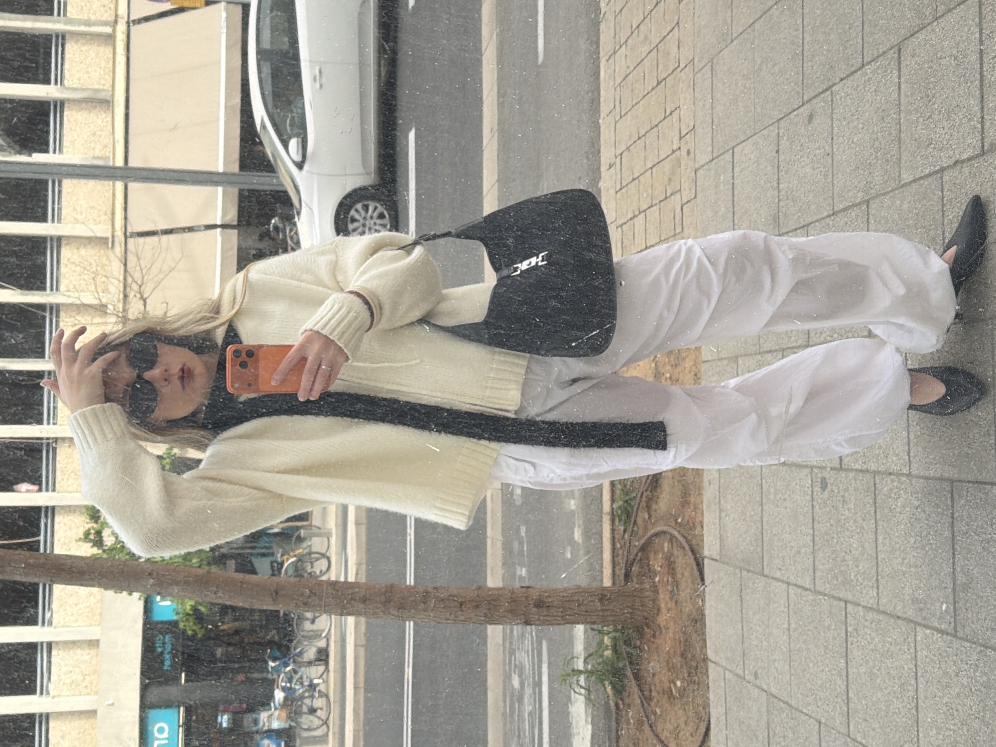 A woman stands on a Tel-Aviv, Israel sidewalk, her image reflected in a storefront window. She wears sunglasses, a cream sweater, white pants, and black flats. A black purse hangs from her shoulder, and she holds an orange phone. The reflection reveals a street scene with a white car, parked bicycles, and buildings.