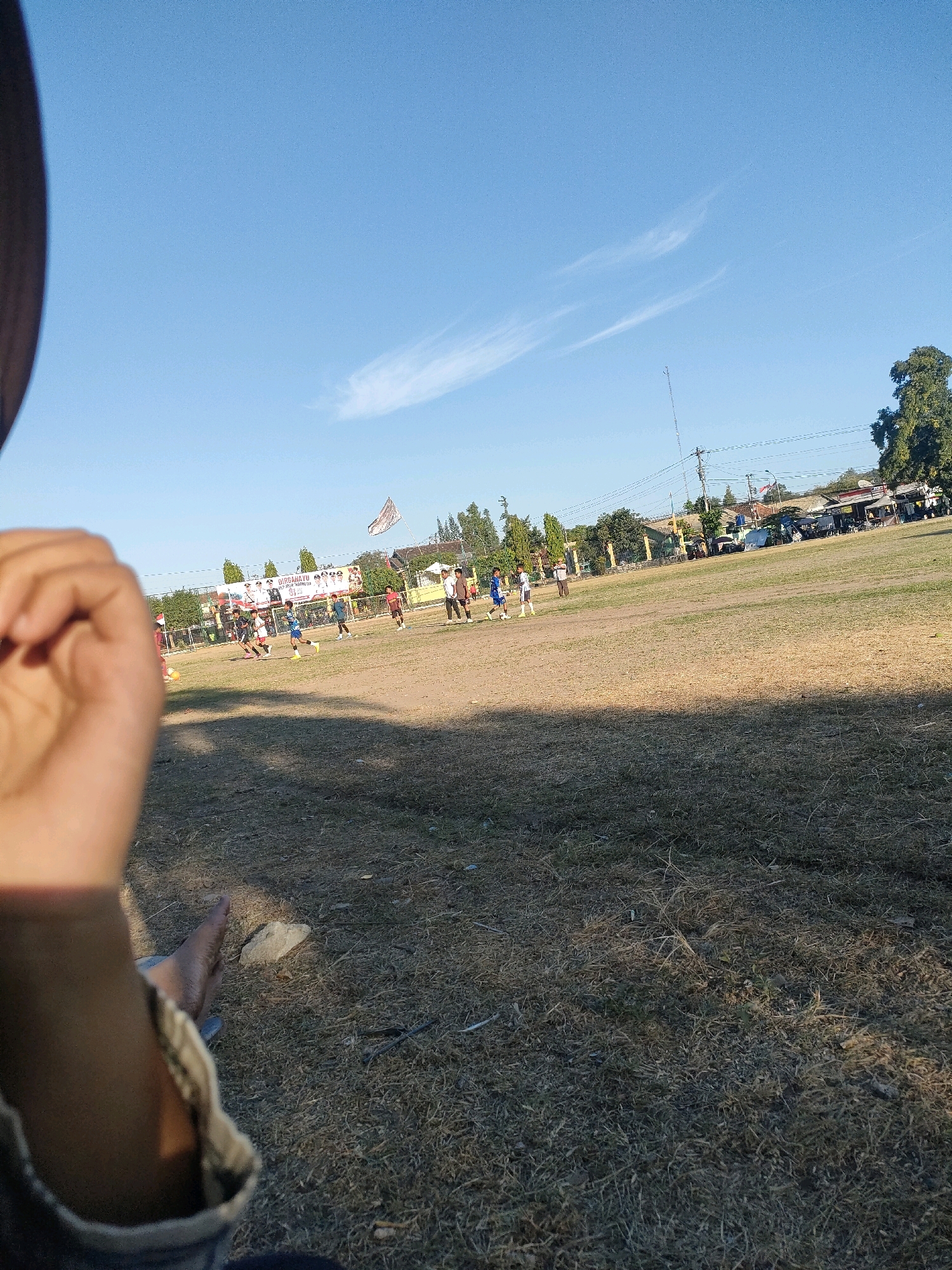 A wide shot of a dusty, dry grass field in Kasihan, Indonesia, shows a group of young boys playing soccer under a bright blue sky with a few wispy clouds. The field is in poor condition, with patches of bare earth and sparse, dry grass. A banner with text in Indonesian hangs in the background, along with a flag on a pole. The boys are scattered across the field, some actively playing the game and others watching. A blurry hand and arm are in the foreground, suggesting the photo was taken by someone sitting or lying on the ground. The scene appears to be an outdoor sporting event or practice in a rural or semi-urban area.