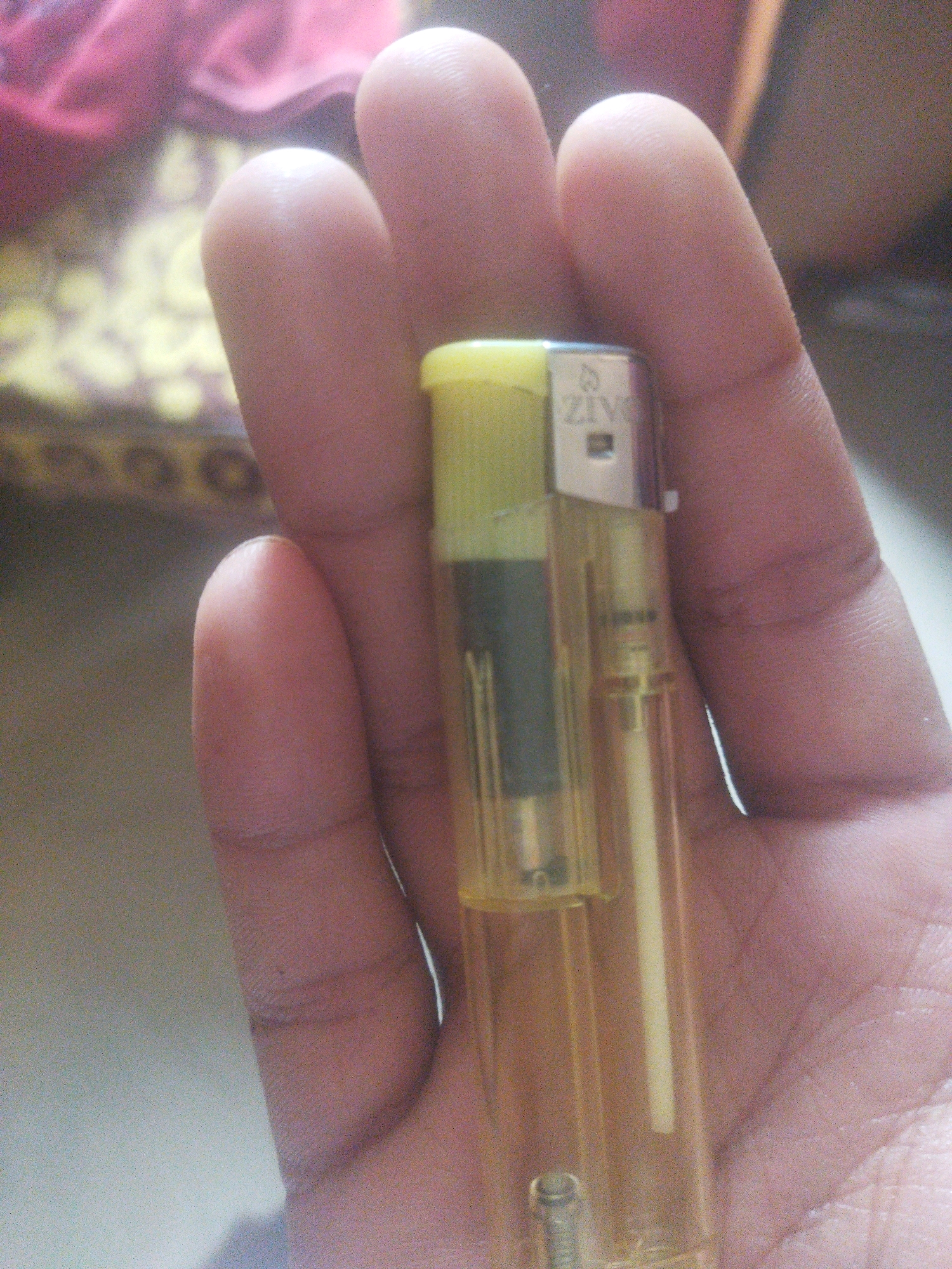 An individual's hand is holding a translucent yellow lighter. The lighter has a yellow cap and a visible fuel reservoir. The brand name "ZIV" is visible on the side of the lighter, along with a flame icon. The background is blurred, showing hints of pink and yellow fabric, suggesting an indoor setting. The lighting is dim.