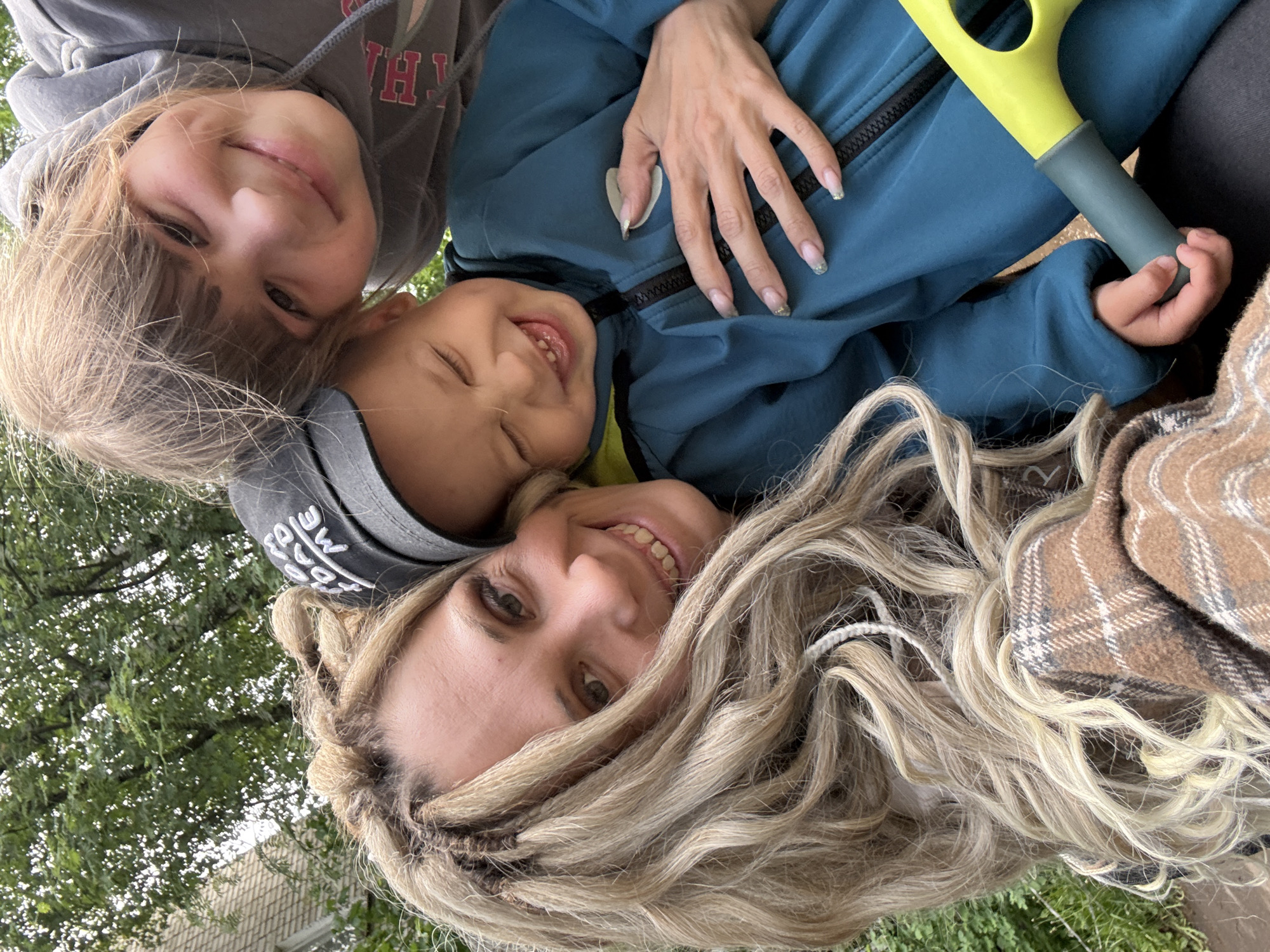 A selfie of three people in Rusanovo, Russia. A woman with long, light-blonde hair in braids and a black baseball cap smiles at the camera. Beside her, a young boy in a dark blue jacket with a yellow inner lining is smiling and winking. To the left of the boy is a young girl with light-brown hair wearing a gray hooded sweatshirt. A person’s hand rests on the boy’s jacket. Part of a green and gray gardening tool is visible. A brown plaid blanket is on the right. Trees and a building are visible in the background.
