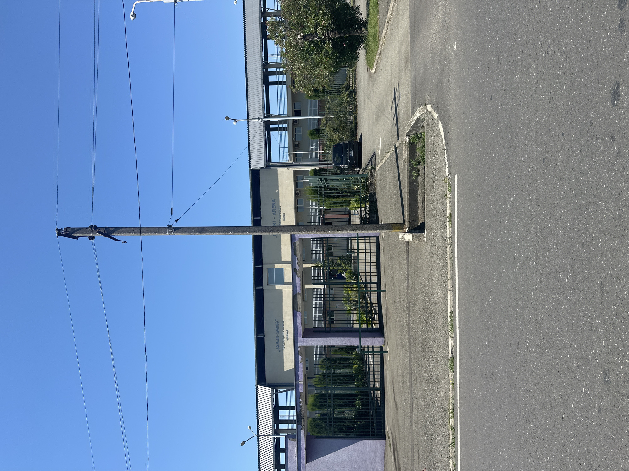 The image depicts the "Poti Arena" located in Poti, Georgia. In the foreground, there is an asphalt road curving to the right, with a concrete curb and a patch of grass along the edge. A concrete sidewalk or pedestrian path is also visible. A utility pole with wires runs vertically in the middle of the frame. Behind the utility pole, a dark gray fence with vertical bars encloses an area with manicured bushes and trees. To the left of the fence, a lavender-colored structure is visible. The main building of the Poti Arena is a long, light-colored structure with a ribbed metal roof extending over a covered walkway. The building has several windows and air conditioning units visible. Text in Georgian and English is present on the building, indicating "POTI ARENA" and other affiliations. A black SUV is parked inside the fenced area. The sky is clear and blue.