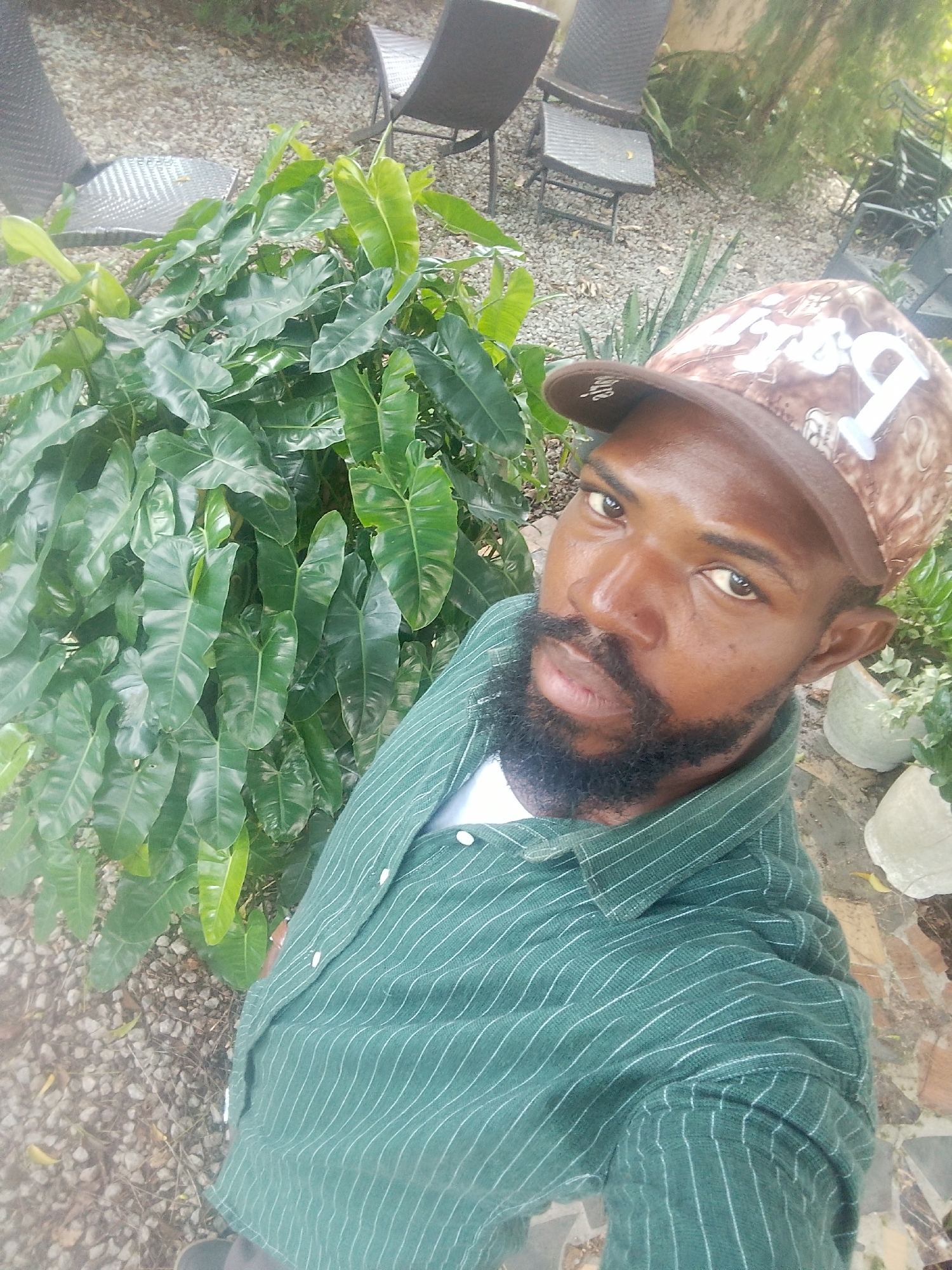 The image shows a close-up, high-angle selfie of a Black man wearing a camouflage baseball cap and a green striped button-down shirt. He has dark skin, a beard, and is looking directly at the camera. The man is standing outdoors, with lush green foliage to his left. The background features gravel, outdoor furniture, and potted plants. The location is identified as Aketu-Oja, Nigeria.