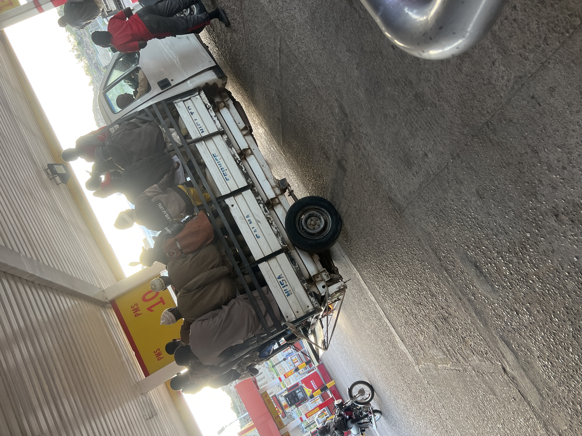 The image depicts a crowded scene at a filling station, possibly in Gura Top, Nigeria. A white utility vehicle with an open back is parked near a gasoline pump. Several people are gathered around the vehicle, with some appearing to be seated or standing in the back. A motorcycle is also parked near the pump. Signs with pricing for PMS (Premium Motor Spirit) are visible. The setting appears to be a busy urban environment with various commercial establishments.
