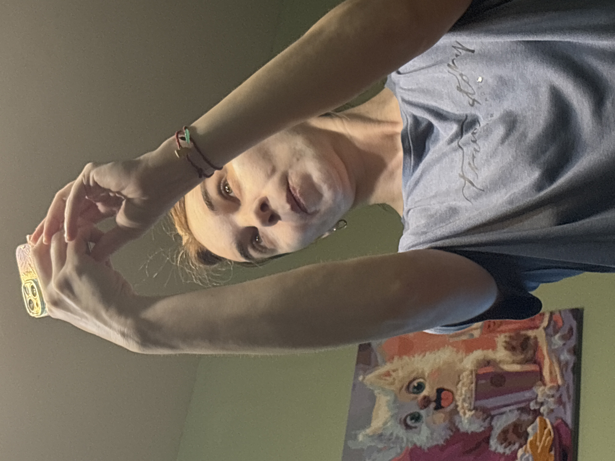 A low-angle, indoor selfie shows a woman holding a smartphone, with her head tilted down. She is light-skinned, with light brown hair partially pulled back. She is wearing a grey t-shirt with writing on it. Her arms are raised to hold the phone, and she is wearing a bracelet on her right wrist.
In the background, a wall is visible with a painting depicting a white cat with a pink background. The setting is unidentified and there is no information on the city of Unknown, Russia.