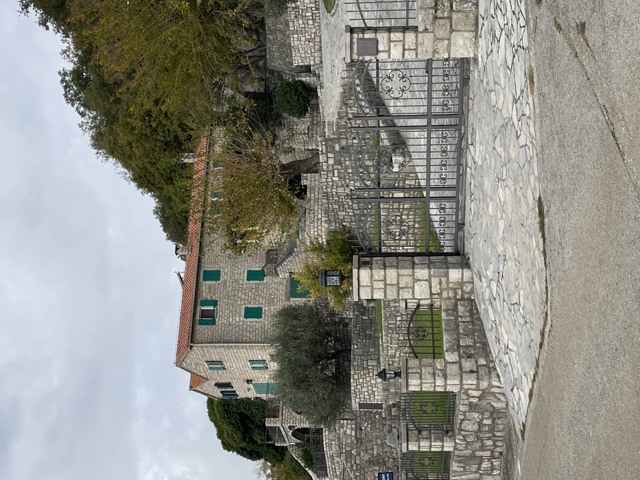 An exterior long shot captures a stone-built residence and its immediate surroundings in Đenjaši Česminovo, Montenegro, under an overcast sky. The multi-story building, constructed of light-colored rough-hewn stone blocks, features a red-tiled gabled roof with two visible chimneys. Numerous rectangular windows are fitted with closed green shutters, while one upper-story window on the main facade displays open green shutters. A dark-railed balcony is visible on the left side of the structure.
The property is situated on a terraced hillside, with extensive retaining walls and staircases built from the same light-colored stone, ascending towards and around the main dwelling. Mature trees, including a prominent olive tree to the left and several deciduous trees exhibiting autumnal foliage, are integrated into the landscaping. Denser green forest covers the higher slopes behind the house, particularly on the right.
In the foreground, a dark asphalt road runs horizontally. A substantial entrance gate, forged from dark metal with decorative scrollwork, blocks access to a driveway paved with irregular light-colored flagstones. This gate is flanked by two robust stone pillars, each capped with a black, lantern-style light fixture. A low stone wall with matching smaller pillars and dark metal fencing extends from these main pillars, defining the property's perimeter. A small, dark rectangular plaque is affixed to the right gate pillar. A partial blue sign is visible on a stone wall behind the left gate pillar. No individuals or active subjects are present in the scene.