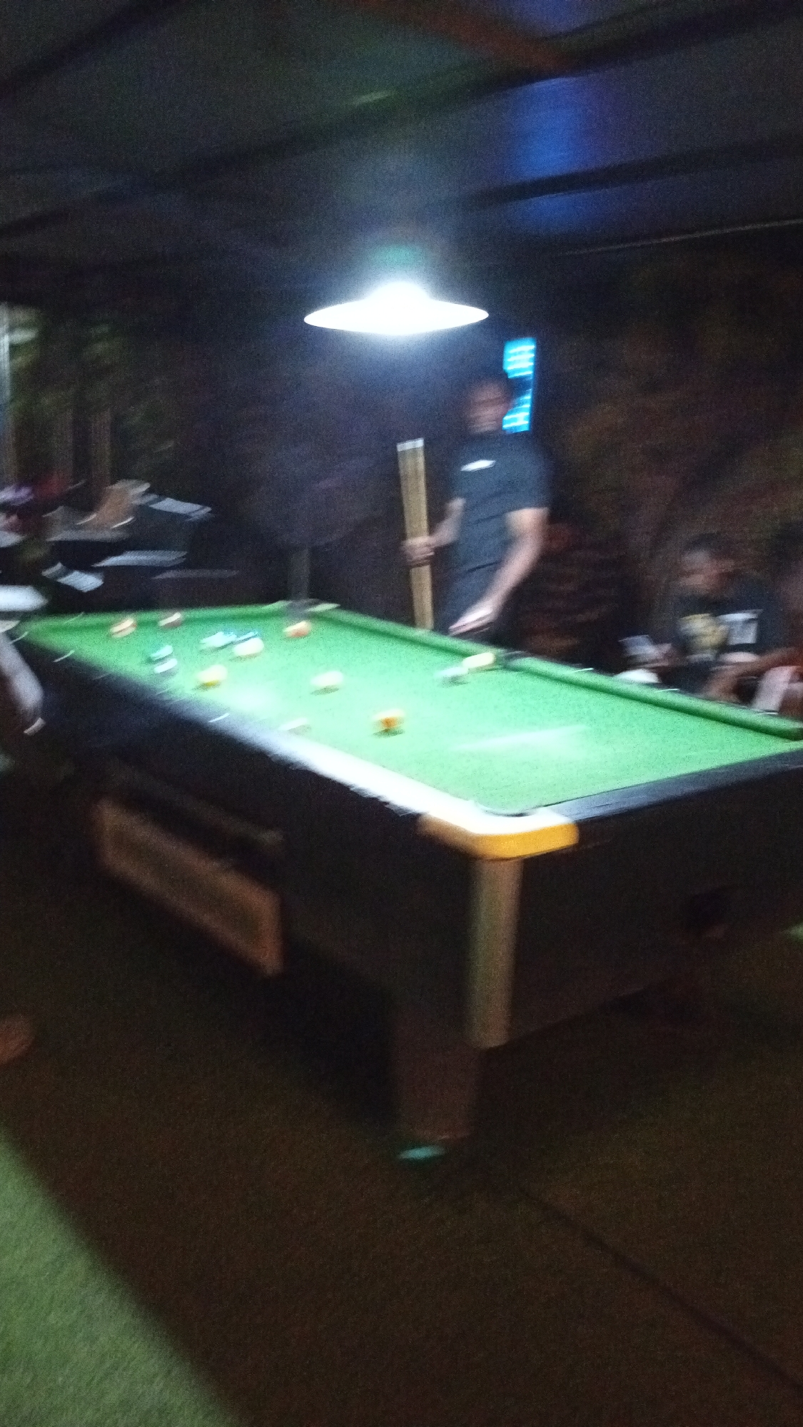 A person in a dark t-shirt is holding a pool cue and standing next to a pool table in what appears to be an indoor establishment. The pool table is set with multiple pool balls. A light fixture hangs above the table. The location of this scene is unspecified, it is not explicitly known that it is located in the city of Adankolo, Nigeria. Other people are present in the blurred background.