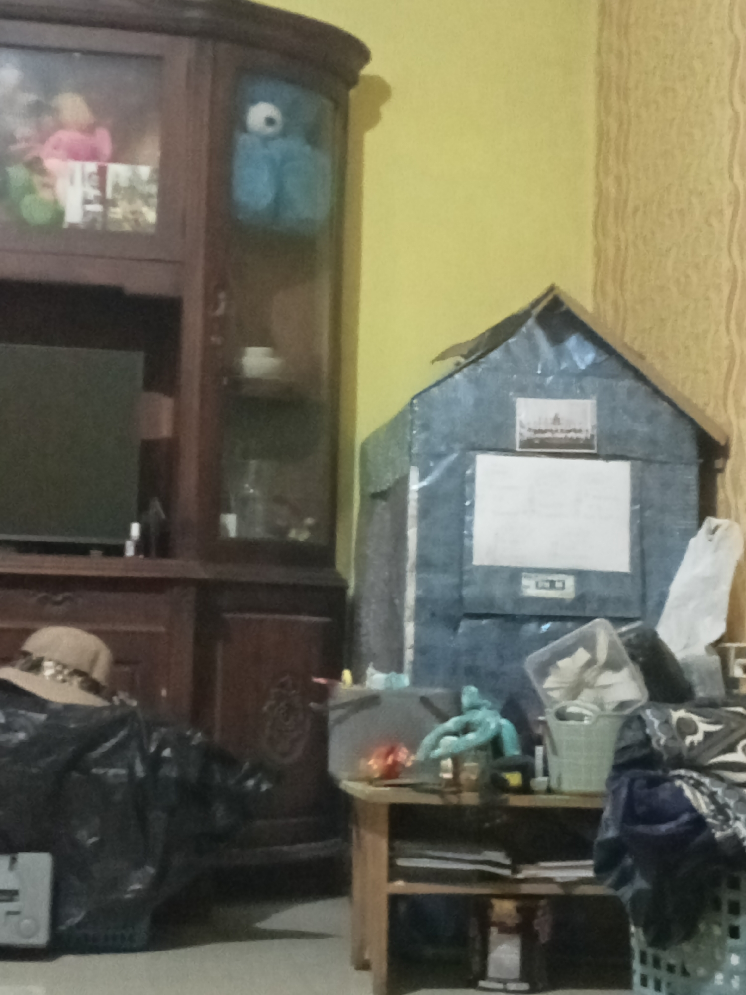 In a room in Grabag, Indonesia, a dimly lit scene unfolds. To the left, a dark wood cabinet with glass doors displays a pink stuffed animal and a blue stuffed animal. Below, a black television screen is visible. In the center foreground, a brown fedora rests on a black tarp covering an unseen object. To the right, a small, makeshift house, constructed from what appears to be blue tarp and cardboard, stands against a yellow, textured wall. A framed picture is affixed to the roof of the structure, and a white paper is attached to its front. A small wooden table sits in front of the makeshift house, holding a clear plastic bin with papers, a green basket, and a crumpled white plastic bag. More items are piled on the table and beside it, including clothing and a green basket in the lower right corner.