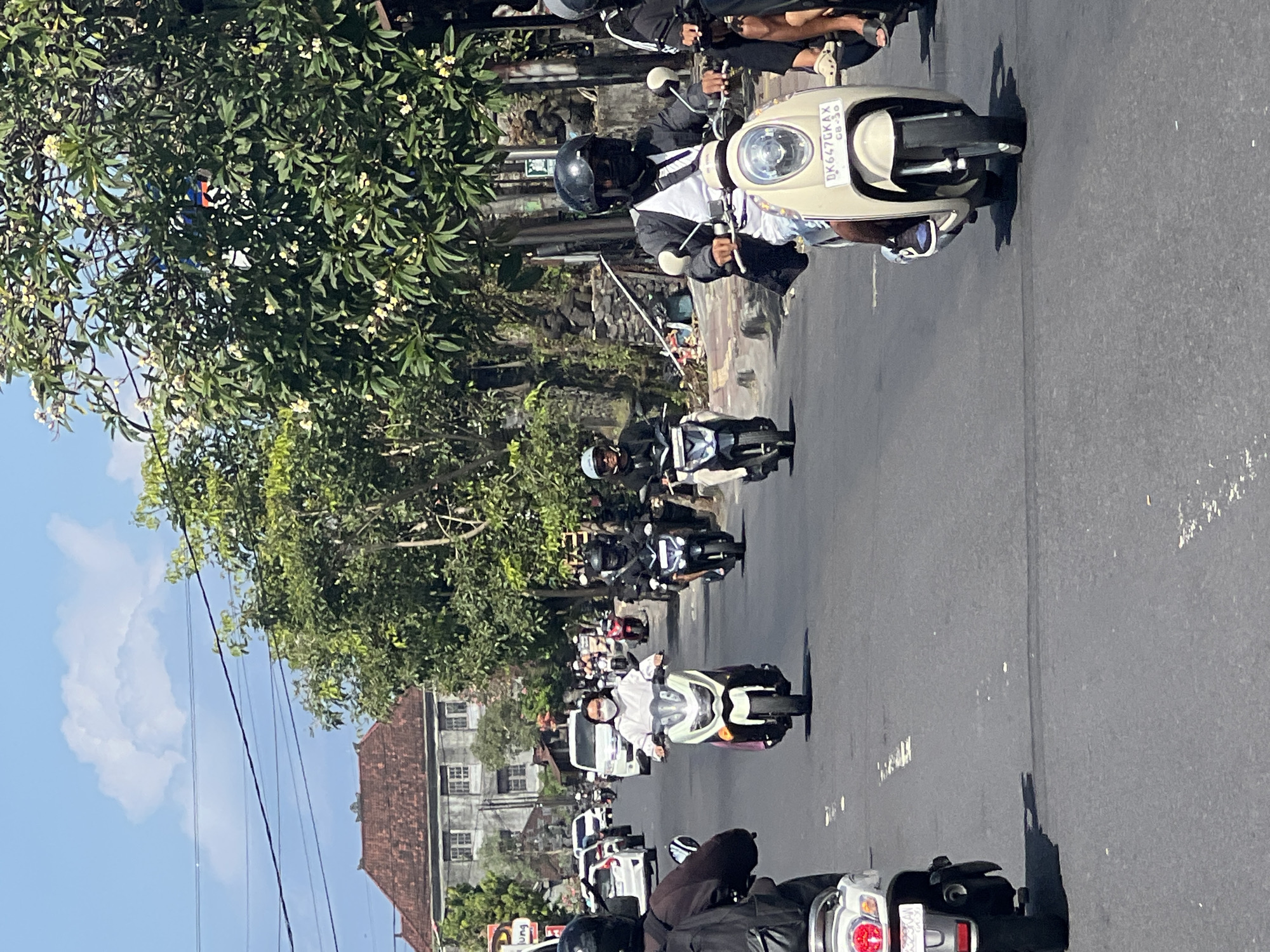 This image depicts a street scene in Sukawati, Indonesia, characterized by a moderate flow of traffic, primarily consisting of motorcycles. The day is sunny, with a clear blue sky and some clouds. The road surface is asphalt, marked with white lines.
Several individuals are seen riding motorcycles, many wearing helmets and some with face masks. In the foreground, on the right side of the road, a person is riding a light-colored scooter, wearing a black helmet and a black and white jacket. On the left side, another person on a motorcycle is partially visible, wearing a black backpack. Further down the road, a woman in a white hoodie and a pink skirt is riding a white and green scooter, also wearing a face mask. Other motorcycles and a few cars are visible in the background, suggesting a typical day in a busy urban or semi-urban area.
The environment includes trees lining the street, with some buildings visible on the left side. One building has a red-tiled roof and traditional Balinese architectural elements. Electrical wires are strung overhead. The overall impression is that of a lively, everyday scene in a tropical climate.