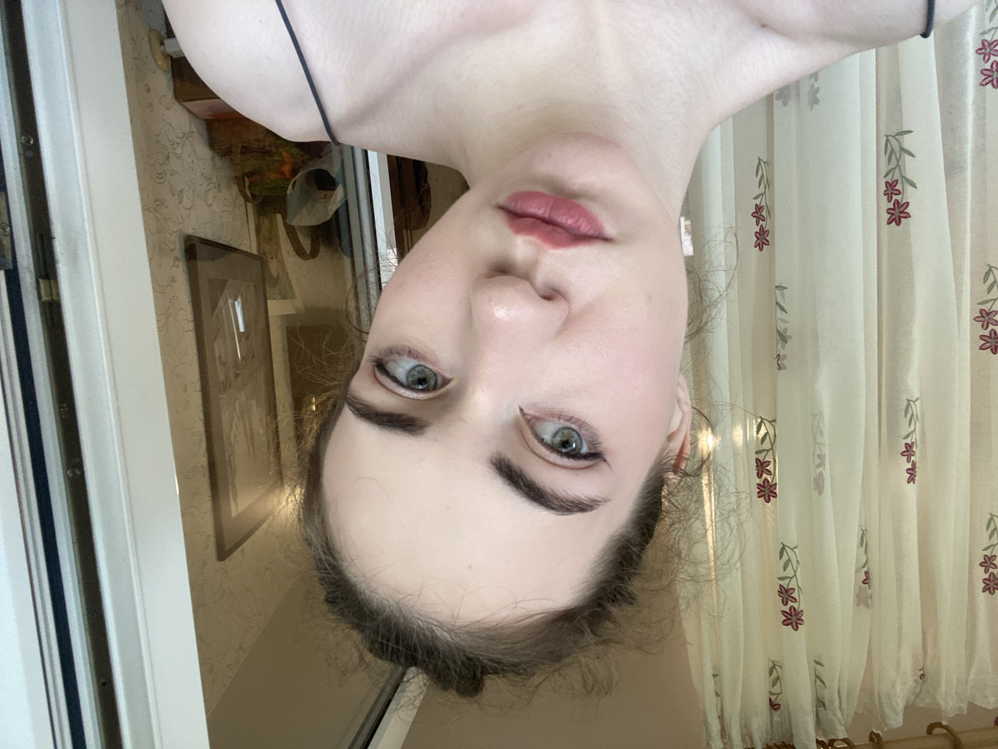 This is a close-up, vertically oriented, upside-down selfie of a person's face, taken indoors. The subject has light skin, green eyes with noticeable eyeliner, and dark, arched eyebrows. Their lips are slightly parted and appear to have a matte lipstick applied. Their hair is dark and appears to be tied back loosely.
The background shows a sheer curtain with a floral pattern of small red flowers and green leaves. To the left of the subject, there is a white door frame and a glimpse of a window with vertical blinds. Further in the background, on the wall, there are framed pictures, suggesting a domestic setting. The lighting appears to be natural, possibly from a window, and the time of day is not explicitly indicated but seems to be daytime. No visible text is present in the image. The overall mood is casual, and the subject's direct gaze towards the camera suggests a moment of self-reflection or informal self-documentation. The location context is Zavodoukovsk, Russia.