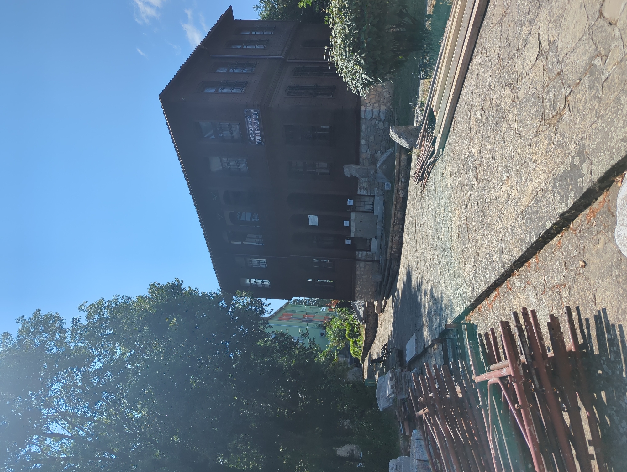 In Malko Tarnovo, Bulgaria, a two-story wooden building with a stone foundation and a red tiled roof stands under a clear blue sky. The building, which appears to be an historical museum, has numerous windows with metal grates and a sign above the entrance. In the foreground, a paved walkway leads towards the building. To the left of the walkway, a pile of red metal scaffolding and construction materials are stacked on the ground. To the right of the walkway, several long pieces of wood are laid out. A large green tree shades a portion of the scene on the left, and a smaller, bushy green tree is situated to the right of the building. In the background, another modern building, painted green and orange, is visible. The overall scene suggests a tranquil, sunny day in a historic town, possibly with ongoing construction or renovation.