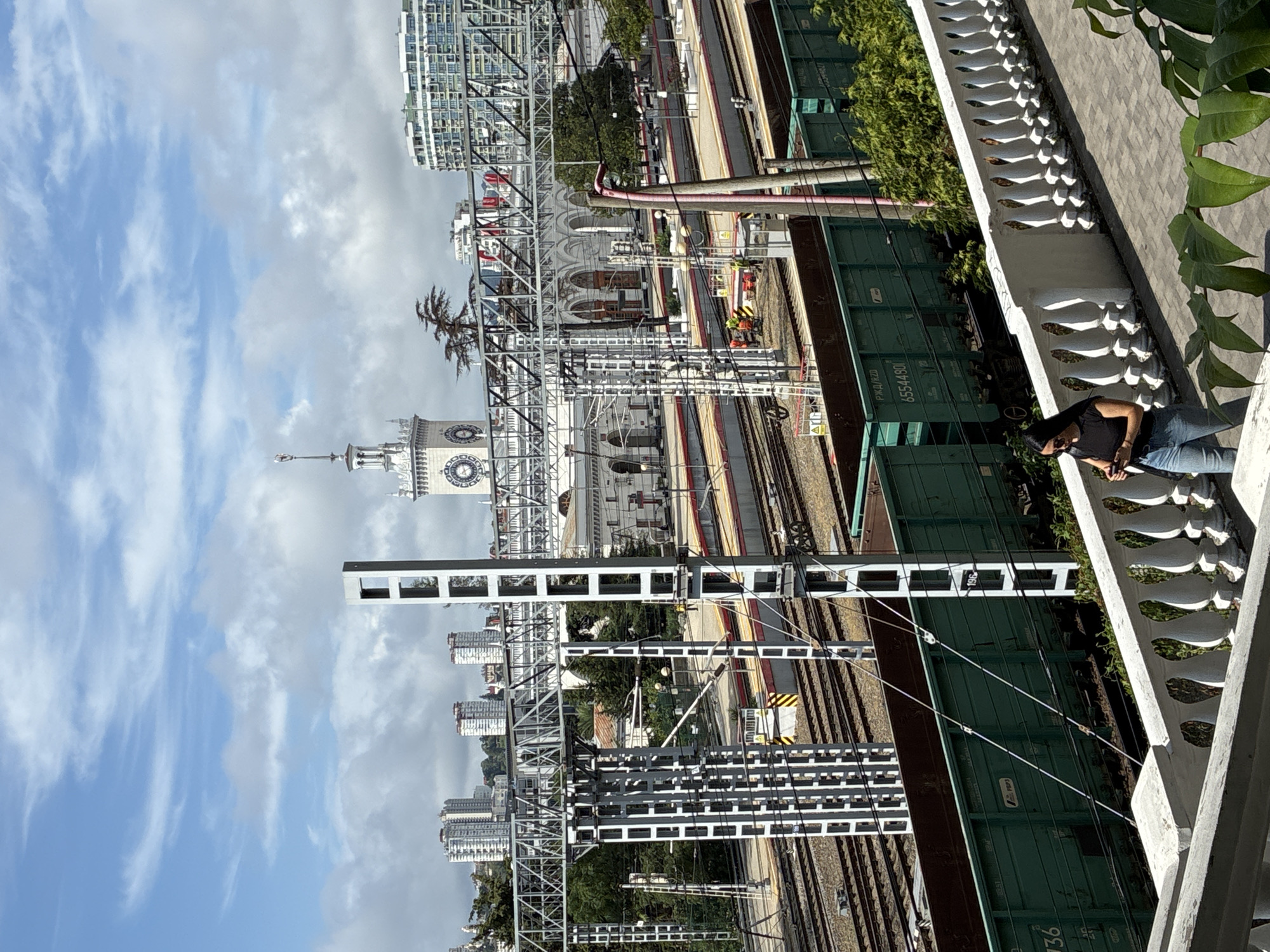 The image shows the Sochi railway station in Sochi, Russia, on a clear day with scattered clouds. A young woman with long dark hair, wearing a black t-shirt and blue jeans, is walking down a paved path, looking at her phone. She is on a balcony with a white ornate railing, with green trees and foliage in the foreground and background.
In the middle ground, there are multiple railway tracks with several green freight trains and passenger trains. Overhead power lines and supporting structures are visible, along with a tall, white clock tower that is characteristic of the station's architecture. In the background, modern high-rise buildings are visible on the hills.
Several railway workers in safety vests are present on the platforms, indicating ongoing operations or maintenance. The overall scene depicts a functional and active railway station in an urban environment.