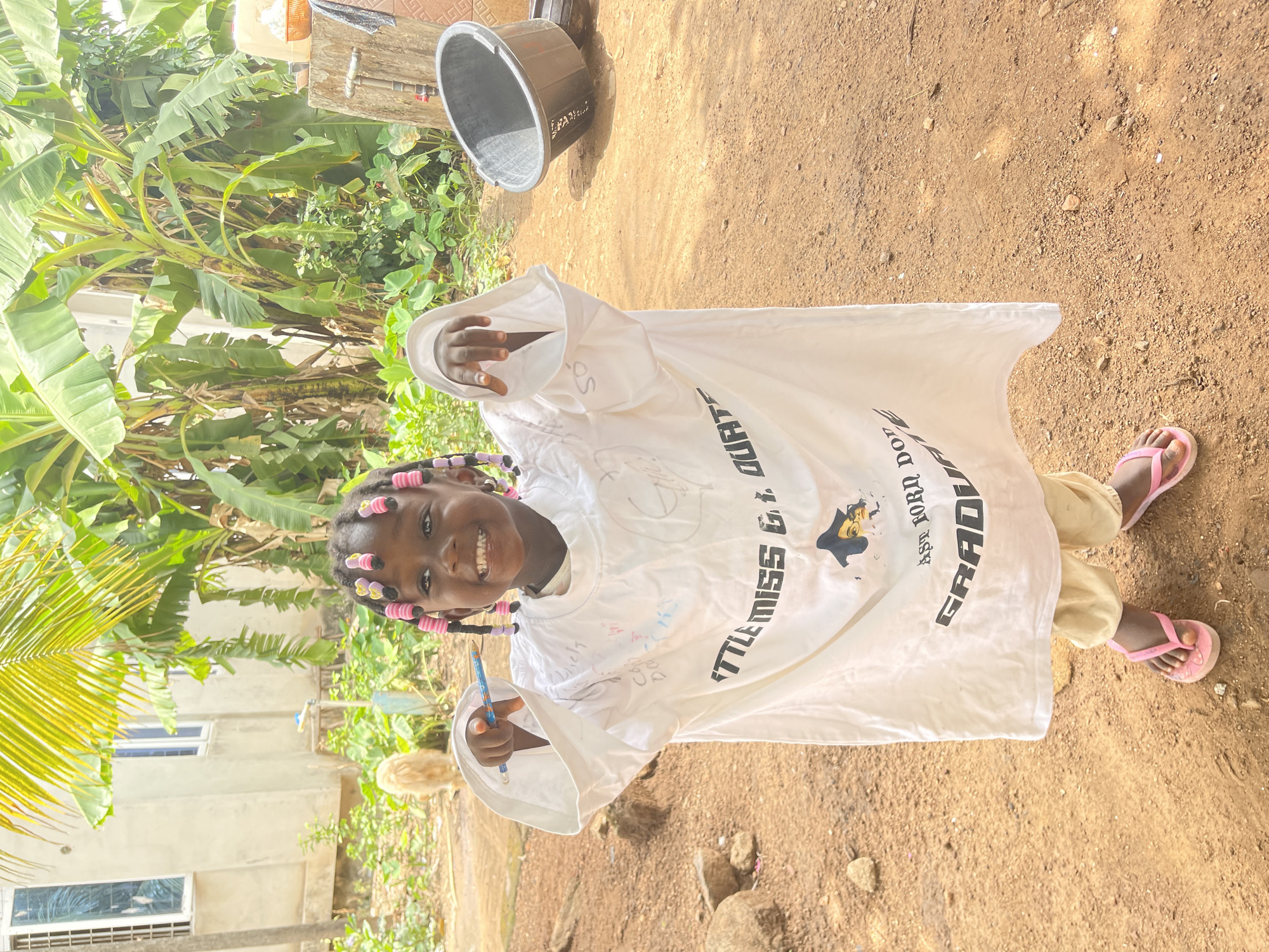 A young girl with dark skin and a broad, happy smile is the central subject of this brightly lit outdoor photograph. Her braided hair is adorned with numerous pink and yellow beads or curlers. She is wearing an oversized white t-shirt that appears to be a graduation shirt, given the visible text, and light-colored pants. On her feet are pink flip-flops. She holds a blue pencil or pen in her right hand, which is held up, as if gesturing or showing the writing on her shirt.
The white t-shirt features printed text, partially visible, including "LITTLE MISS G.T. QUART" (with "QUART" clearly legible) and "GRADUATE," along with "EST. 2024." Additionally, the shirt is covered in various handwritten signatures and messages, indicating it may be a commemorative item.
The setting appears to be a sunny outdoor area, likely during the day. The ground is composed of reddish-brown dirt, with small stones scattered across it. In the background, there is abundant green foliage, including large banana tree leaves and other tropical plants, suggesting a lush environment. A glimpse of a light-colored building with windows is visible behind the greenery. A black plastic bucket rests on the ground to the right of the girl.
The overall scene conveys a cheerful and celebratory mood, possibly marking a school event like a graduation in Akwanga, Nigeria, a location known for its natural vegetation and warm climate.