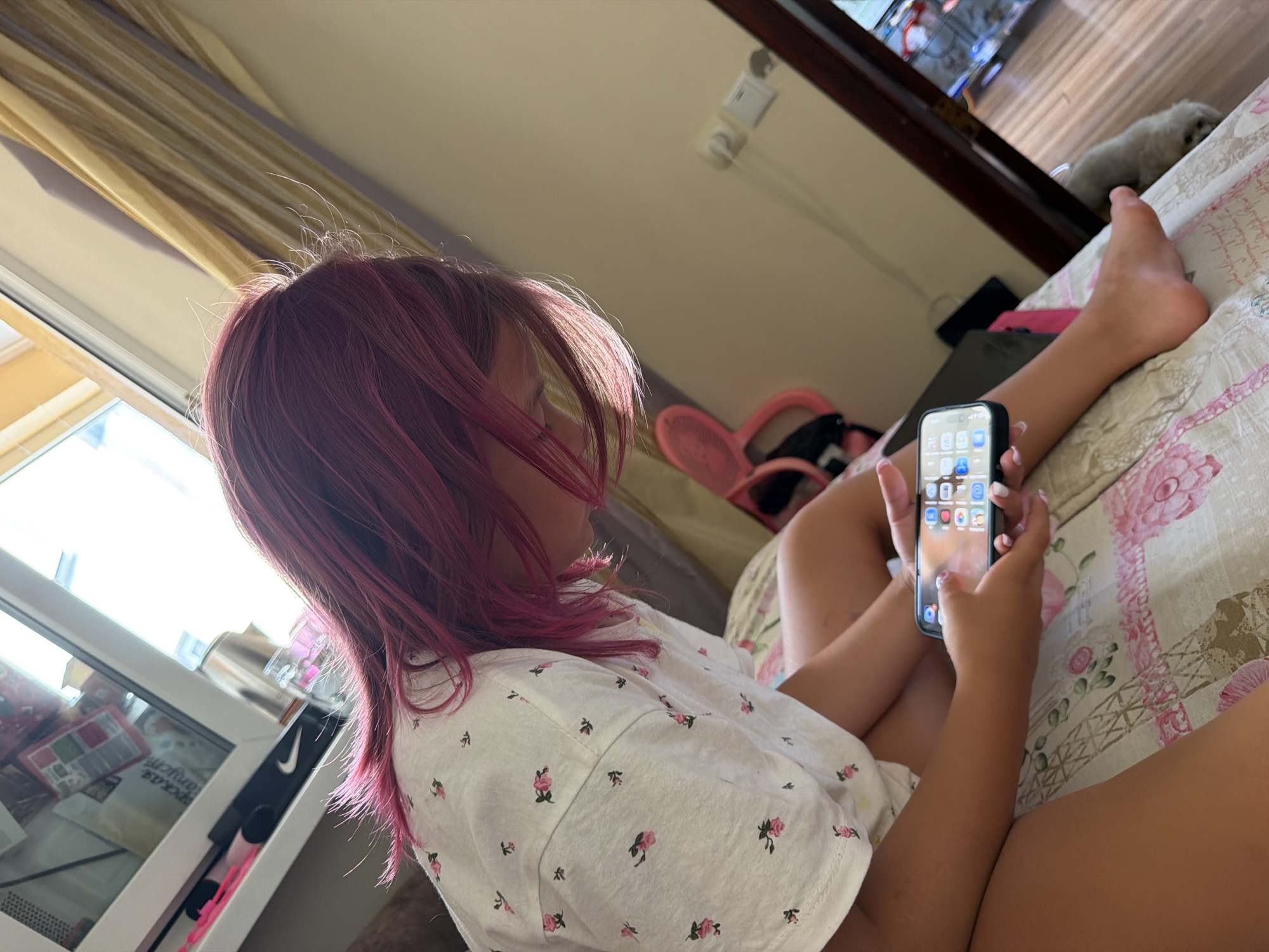 The image depicts a young girl with vibrant pink hair sitting on a bed, looking down at her smartphone. She is wearing a white t-shirt adorned with a floral pattern. The bedspread features a large print of the Eiffel Tower. Sunlight streams in from a nearby window, illuminating the scene. The girl's legs and feet are visible, and her hands are engaged with the device. In the background, a pink hair dryer and a mirror reflecting a room with wooden floors and electrical outlets are observable. A small, light-colored dog is partially visible near the mirror. The scene appears to be set indoors, likely in a residential area. There is no specific information within the image that allows for determination of the location within the city of Partenit, Ukraine.
