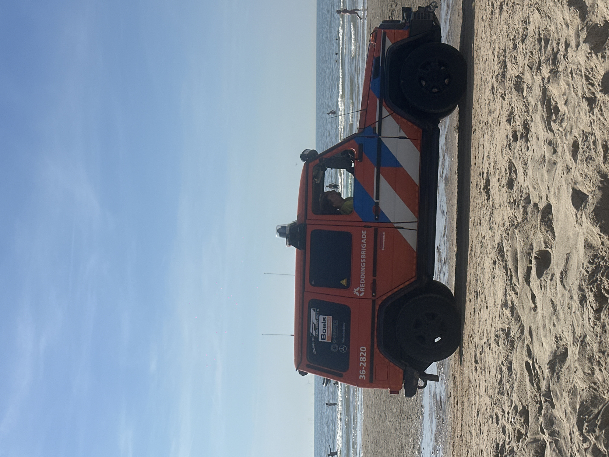 An orange Mercedes-Benz G-Class vehicle, marked with "REDDINGSBRIGADE" and the number "36-2820", is parked on a sandy beach in Zandvoort, Netherlands. The vehicle is equipped with a light bar on the roof and has red, white, and blue diagonal stripes along its side. A person is visible in the driver's seat, looking out the window. In the background, the sea is calm with gentle waves, and several people are visible in the water, some swimming and others standing. The sky is clear and blue with a few wispy clouds. The foreground consists of rippled sand, indicating a typical beach environment.