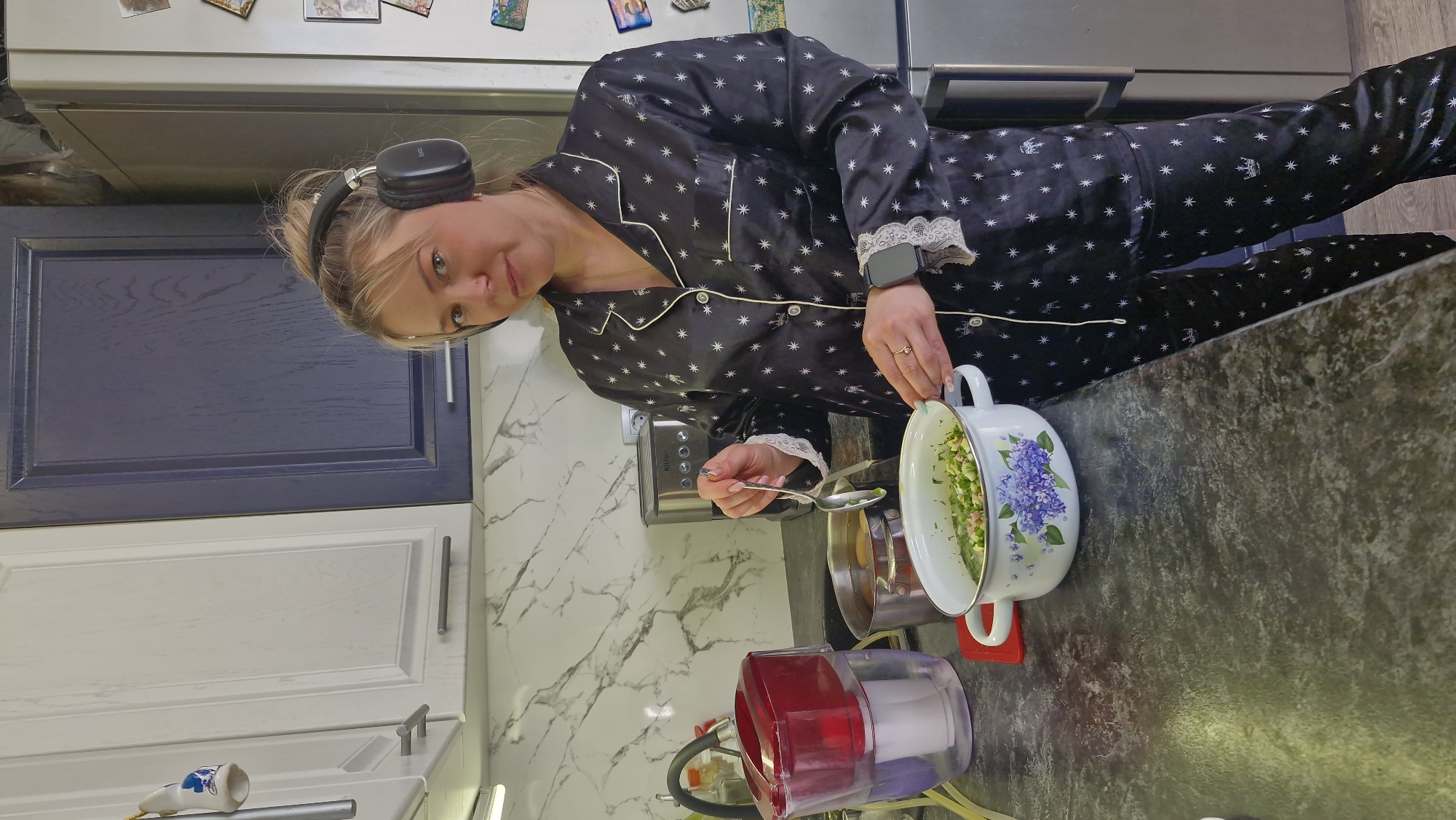 The image, rotated 90 degrees clockwise, depicts a woman in a kitchen in Otchor, Russia, engaged in food preparation. She has blonde hair, possibly tied back, and is wearing black over-ear headphones with "bt" faintly visible on one ear cup. She is dressed in black satin pajamas adorned with white stars and featuring white piping and lace trim on the cuffs. A dark-colored smart watch is on her left wrist, and a ring is visible on her right hand. She looks directly at the viewer with a calm expression.
In her right hand, she holds a spoon, while her left hand is near a white enamel pot with a purple floral design, which contains chopped green ingredients, likely a salad or other food being prepared. The pot rests on a dark grey/black countertop, alongside a red and purple water filter pitcher. Behind her, there's a white marble-patterned backsplash and a silver toaster or small kitchen appliance with the brand name "Kitfort" clearly visible on its side.
The kitchen features white lower cabinets and navy blue upper cabinets, all with modern silver handles. A stainless steel refrigerator, decorated with various magnets, is visible in the background. The overall scene suggests a casual, domestic moment, possibly in the morning or evening given her attire. The lighting appears to be artificial and bright.