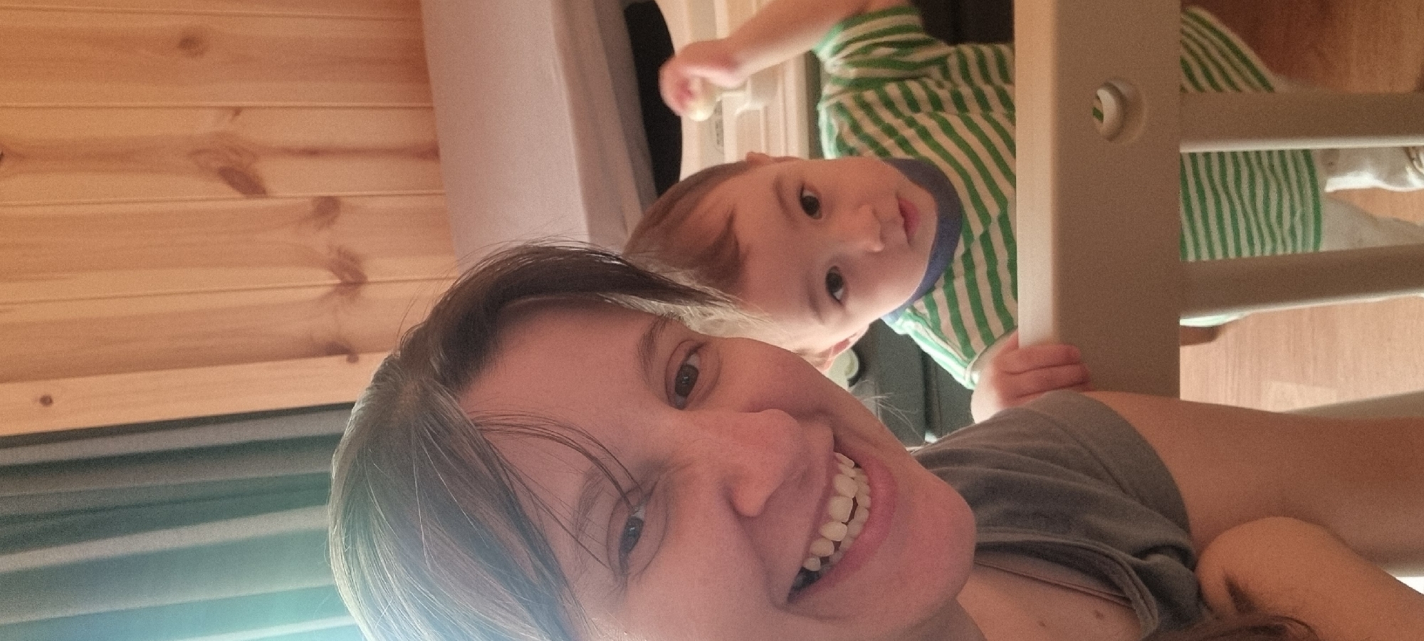 A woman and a child are visible in a photograph. The woman, with dark hair and fair skin, is smiling at the camera. A child wearing a green and white striped shirt is positioned above the woman, partially obscured by what appears to be a light-colored wooden structure with horizontal slats. The background includes a wood-paneled wall and a neutral-colored surface.