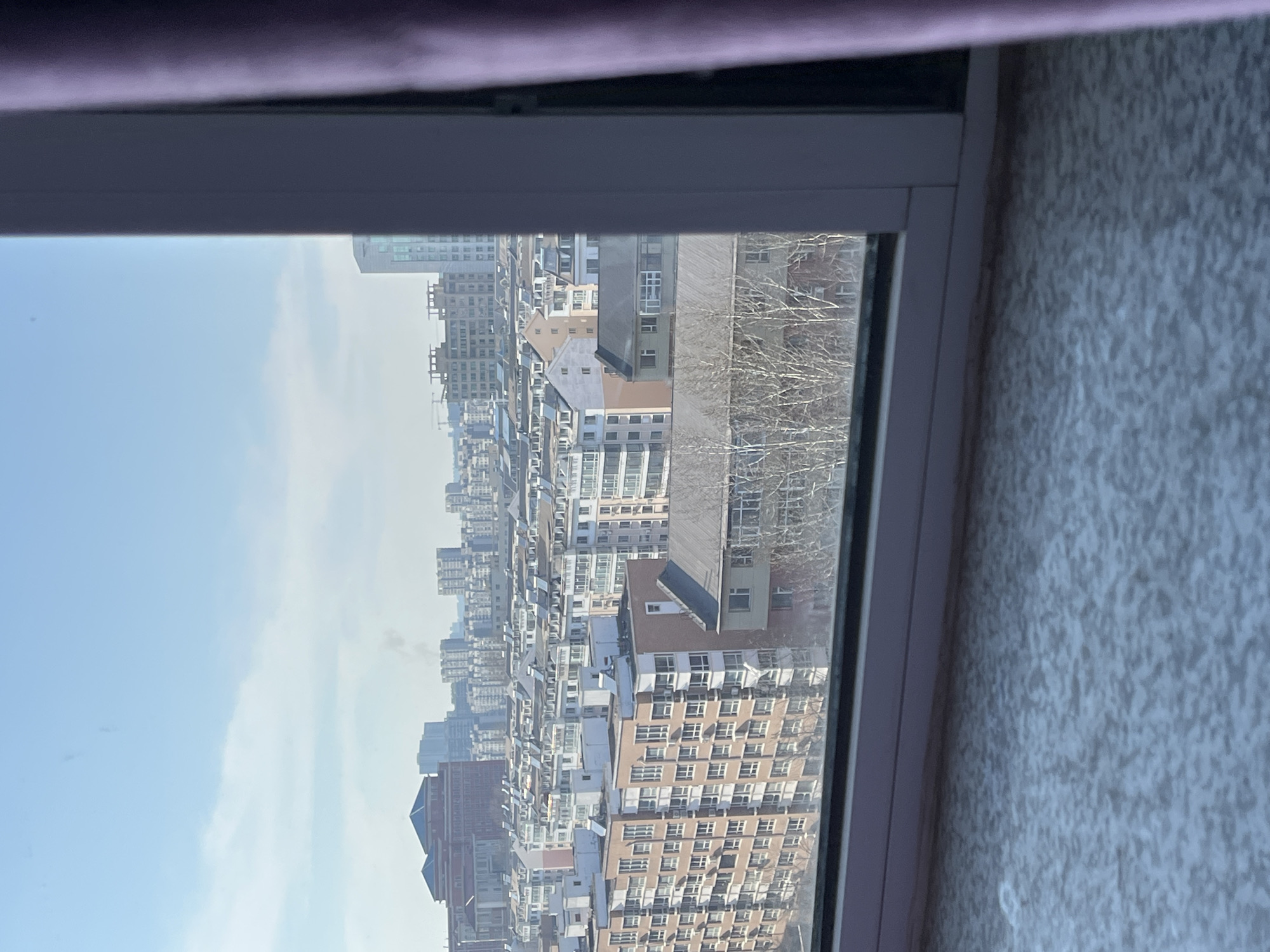 The image depicts a dense urban landscape in Changchun City, China, as viewed through a window. The scene showcases numerous high-rise apartment buildings, tightly packed together and characterized by a mix of architectural styles and colors, predominantly in shades of white, grey, and brown. The sky is visible in the background, with a pale blue hue and some cloud cover. Bare tree branches are seen through the window in the middle of the image, indicating that the season is either late autumn or winter. The buildings appear to be residential apartments based on the window arrangements. The viewpoint is likely from an upper floor of a high-rise building. No people, subjects, actions, or interactions are observed in the scene. The environment is a typical crowded urban environment common in Chinese cities.