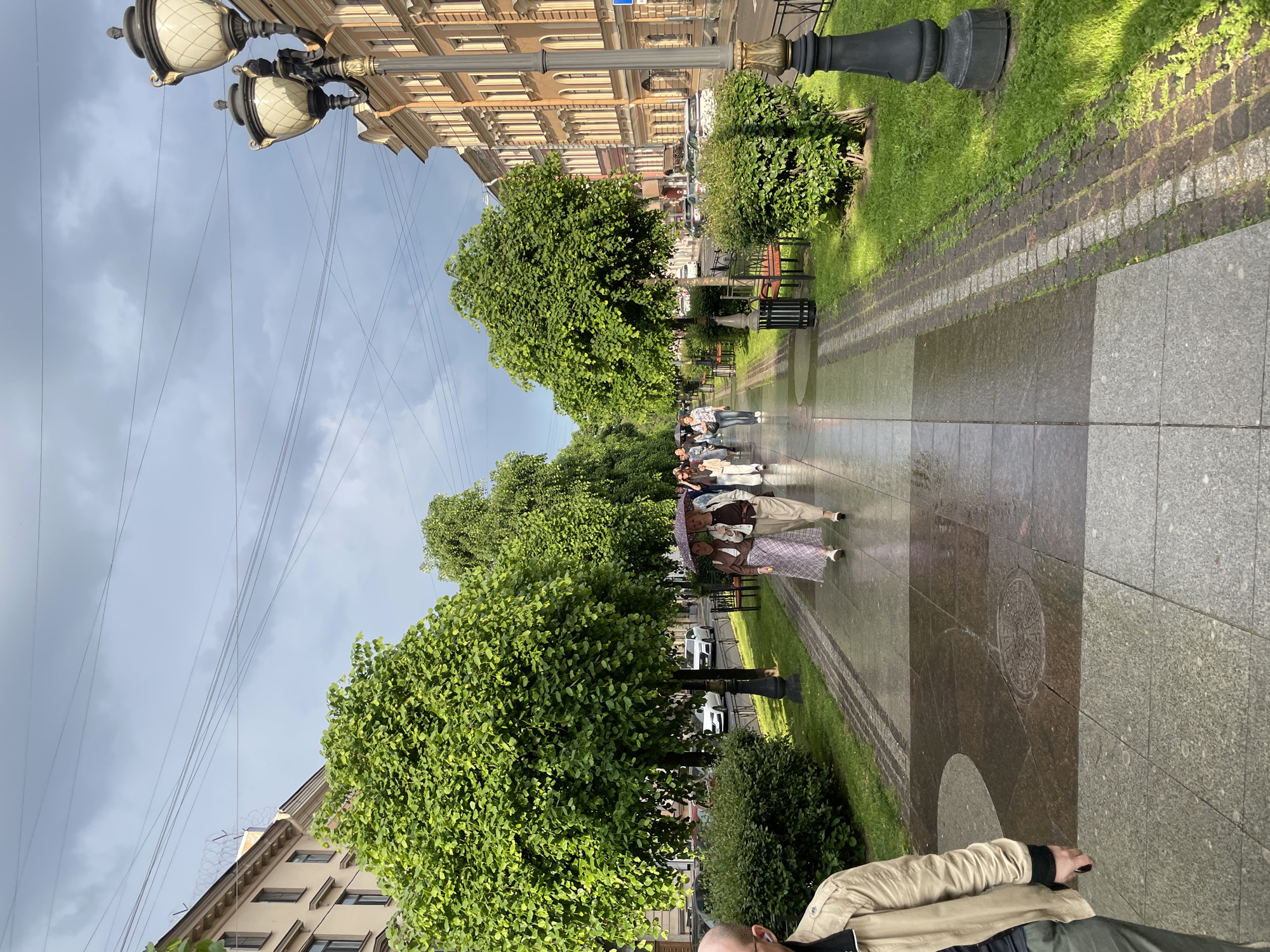 The image, rotated 90 degrees counter-clockwise, depicts a wide urban promenade or avenue, likely in Saint Petersburg, Russia. The scene is captured on an overcast day, evident from the cloudy sky and the visibly wet, reflective pavement, suggesting recent rainfall.
In the foreground, the upper part of a person wearing a beige jacket is visible on the right. The pedestrian path is paved with large, rectangular granite tiles, alternating between darker and lighter shades, creating a patterned surface. A circular, ornate manhole cover is visible on the path.
The middle ground features a long, tree-lined avenue with lush green trees forming a canopy over parts of the path. To the left of the pedestrian area, there's a strip of green grass interspersed with bushes and black park benches. To the right, several ornate streetlights with multiple white, patterned globes rise from black fluted poles, extending along the path into the distance. A group of people is walking along the wet pavement; one person holds an umbrella, reinforcing the recent rain. Other figures can be seen further down the avenue.
In the background, beyond the trees and streetlights, classic, multi-story European-style buildings with numerous windows and detailed architectural facades line the street. A glimpse of a road with parked cars is visible between the trees and buildings. The sky above is largely cloudy, with numerous utility wires crisscrossing overhead.
The overall impression is one of a busy but pleasant urban setting, with people engaged in a casual stroll despite the damp weather. No discernible text is visible in the image.