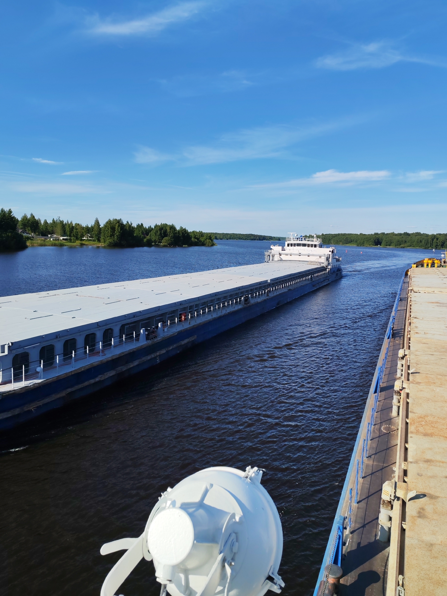 The image depicts a serene scene on a wide, dark blue river under a bright, clear blue sky dotted with scattered white clouds. The sun is high in the sky, indicating it is likely midday. The weather appears to be warm and pleasant.
The main subjects are two large barges navigating the river. The barge in the foreground, viewed from its deck, is a substantial vessel with a long, flat, light gray deck. Its blue hull is partially visible along the side. In the mid-ground, another barge is moving away from the camera, also with a long, flat, light gray deck. This barge has a white superstructure at its rear, which includes cabins and navigation equipment. The dark blue water of the river reflects the bright sky and the surrounding greenery.
The riverbanks are lined with dense, green trees, creating a lush natural backdrop. On the left bank, some houses and smaller structures are visible nestled among the trees, suggesting a populated area along the river. The barges are moving in the same direction, implying they are part of a convoy or traveling along the same route. The overall atmosphere is peaceful and tranquil, conveying a sense of calm movement and natural beauty. The location is identified as Nova, Russia, further specifying the setting. There is no visible text in the image.