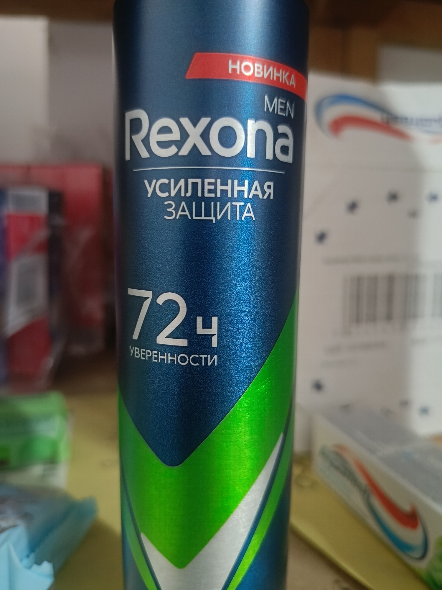 This media file is a close-up photograph of a blue aerosol can of Rexona Men deodorant. The can features prominent white text that reads "Rexona," "MEN," and "УСИЛЕННАЯ ЗАЩИТА" which translates to "Enhanced Protection." Below this, larger white text displays "72Ч УВЕРЕННОСТИ" meaning "72H Confidence." A red label at the top of the can reads "НОВИНКА," which means "New Arrival" or "New Product." The can has a metallic sheen and a diagonal green and white graphic design.
The background is blurred, but it appears to be an indoor setting, possibly a shelf or table, with other objects out of focus. To the right, a blurred white box with a red and blue curved design is visible, possibly another personal care product like toothpaste. To the left, partially visible objects suggest a retail or home environment.
The lighting suggests it is daytime, and the overall impression is that of a product shot or a casual photograph taken of an item in its environment. There are no people visible, and the focus is solely on the deodorant can. The setting is likely a store shelf or a home counter, given the presence of other products. The image does not provide any information about the weather or specific time of day beyond daylight. The location context provided is Berdiansk, Ukraine.