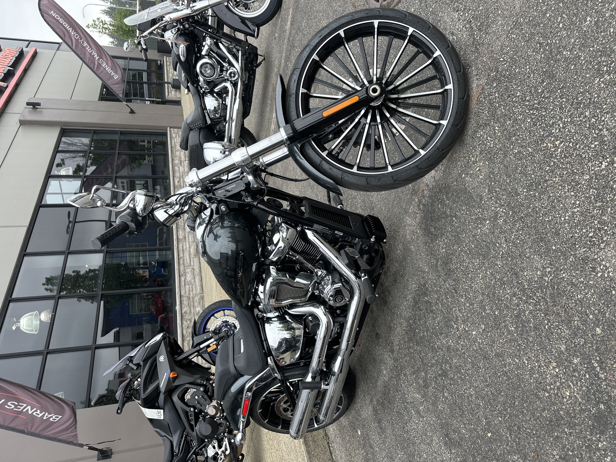 This image captures a close-up view of a black Harley-Davidson motorcycle parked on an asphalt surface outside a building. The motorcycle is the primary subject, showcasing its chrome details, black paint, and a large front wheel with a distinctive black and silver spoke design. A second, partially visible motorcycle is parked behind it.
The setting appears to be a dealership, indicated by the large glass windows of the building and a banner in the background that reads "BARNES HARLEY-DAVIDSON". The building has a modern facade with gray siding and stone accents. The time of day is difficult to determine precisely, but the lighting suggests it could be daytime, possibly overcast given the somewhat muted reflections in the windows. No people are visible in the image. The overall mood is still and commercial, focusing on the display of the motorcycles.