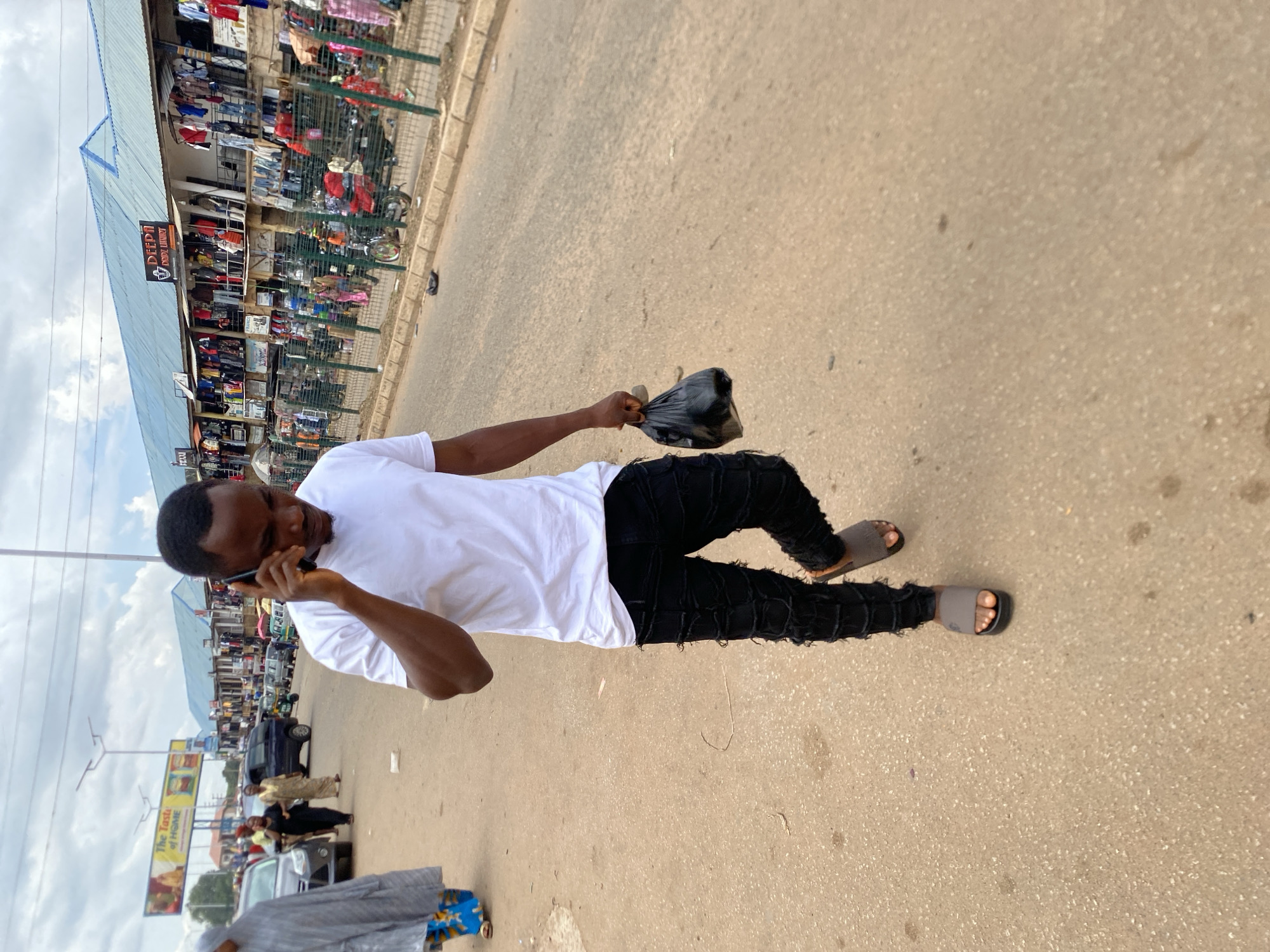 A young adult male, of dark complexion, is depicted walking on a paved road under an overcast sky in what appears to be a market area of Bukuru, Nigeria. He is wearing a white short-sleeved t-shirt, black distressed jeans, and grey slide sandals. He holds a black smartphone to his right ear with his right hand, seemingly engaged in a conversation, while his left hand carries a small black plastic bag. His gaze is directed downwards and slightly forward.
In the background, to the left, a person clad in a grey hijab and a blue patterned long skirt is partially visible, walking in the same direction. Further down the road, several individuals and vehicles, including cars and an auto-rickshaw, are present, indicating a busy street scene. On the right side of the road, a long, multi-story building with a light blue roof houses numerous market stalls or shops, identified by signs such as "DEEPN," "DRODZ LUXURY," "FEAXJ," and "FRANCE." Merchandise, primarily clothing, hangs displayed outside these establishments. A green metal fence separates the pedestrian area from the shops.
An advertising billboard, partially visible on the left side of the road, features the text "The Taste of HOME" and shows images of food items. The road surface is light grey asphalt or concrete, with some minor debris and discoloration. The overall scene suggests an active commercial district.