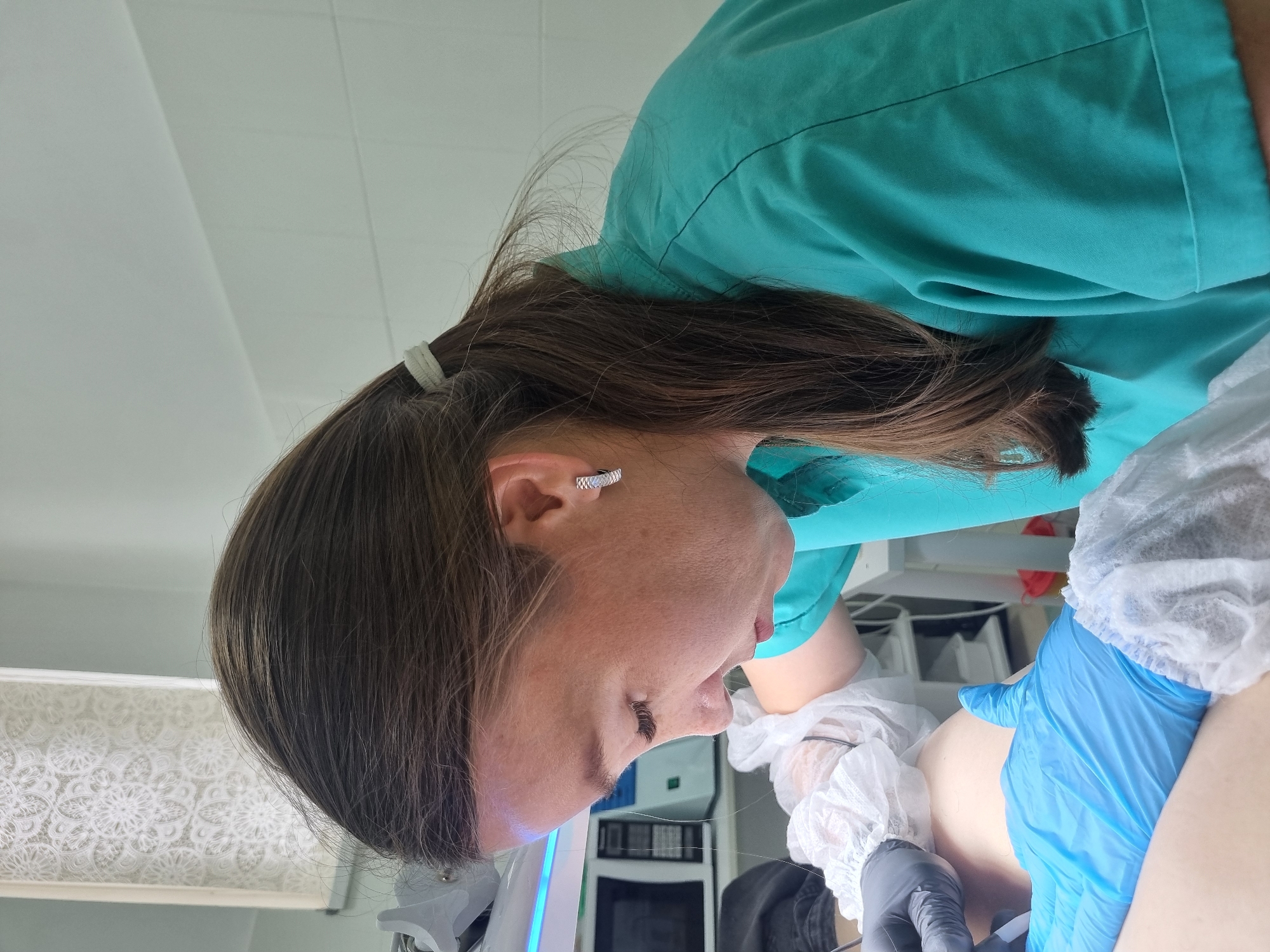 The image shows a close-up of a person, likely a medical professional, wearing a teal scrub top and blue gloves, performing a procedure on another person who is out of focus. The person in the foreground has long brown hair tied back with a light-colored band and is wearing a silver hoop earring. The background shows a white tiled wall and some medical equipment, including what appears to be a monitor with a blue light. The setting is indoors, possibly a clinic or hospital room in Chaikovsky, Russia.