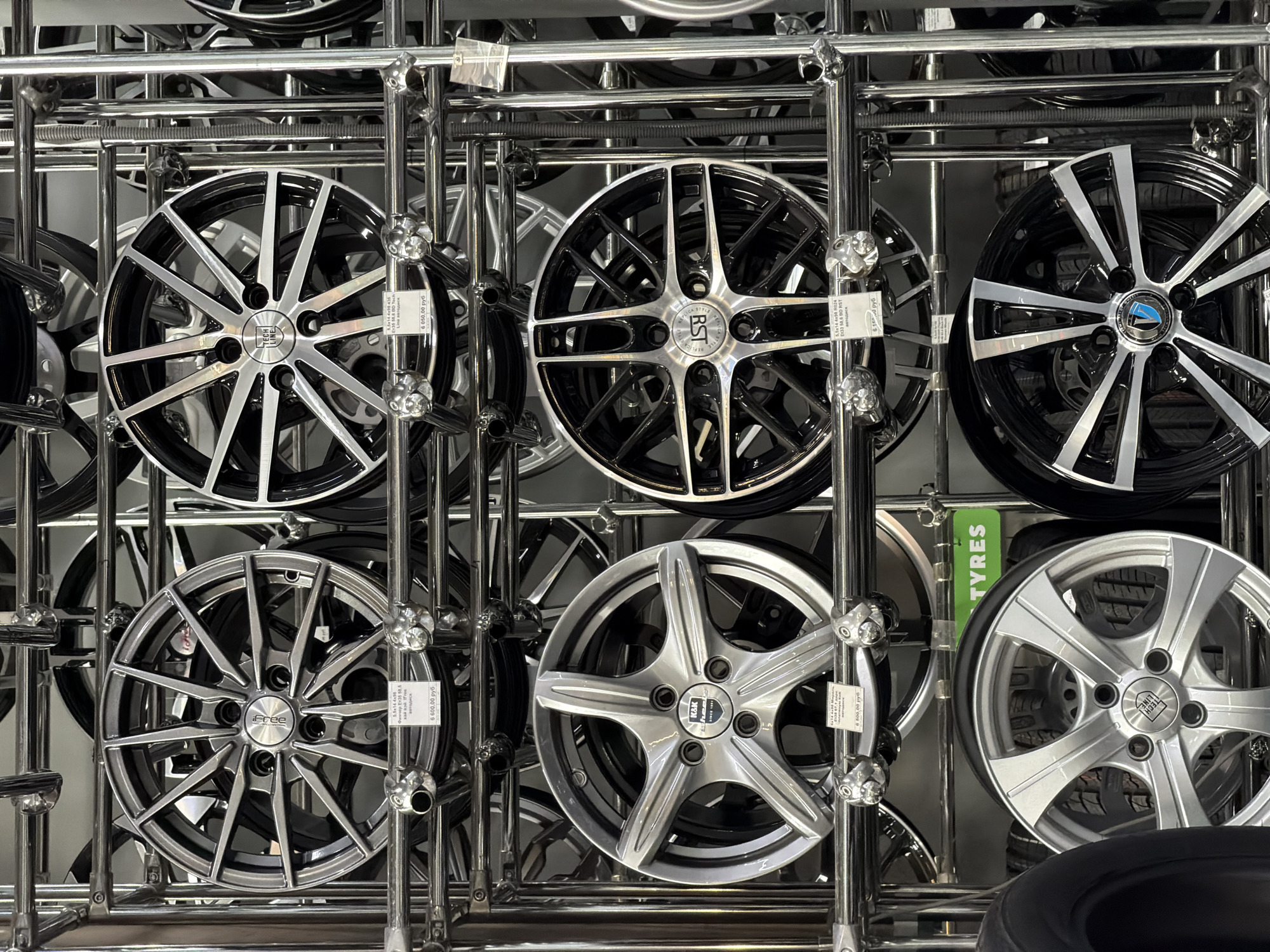 The image shows a close-up of a metal rack filled with car wheels or rims in a store. The wheels are of various designs, with silver and black finishes predominating. Some have brand logos such as "Tech Line", "RST Replica Style", "K&K", and "iFree" visible on their center caps. Price tags in Russian currency (руб) are attached to some of the wheels. A green sign with the word "TYRES" is partially visible in the middle-right section of the image. Based on the extracted text on the tags and the wheels, the location is Izhevsk, Russia.