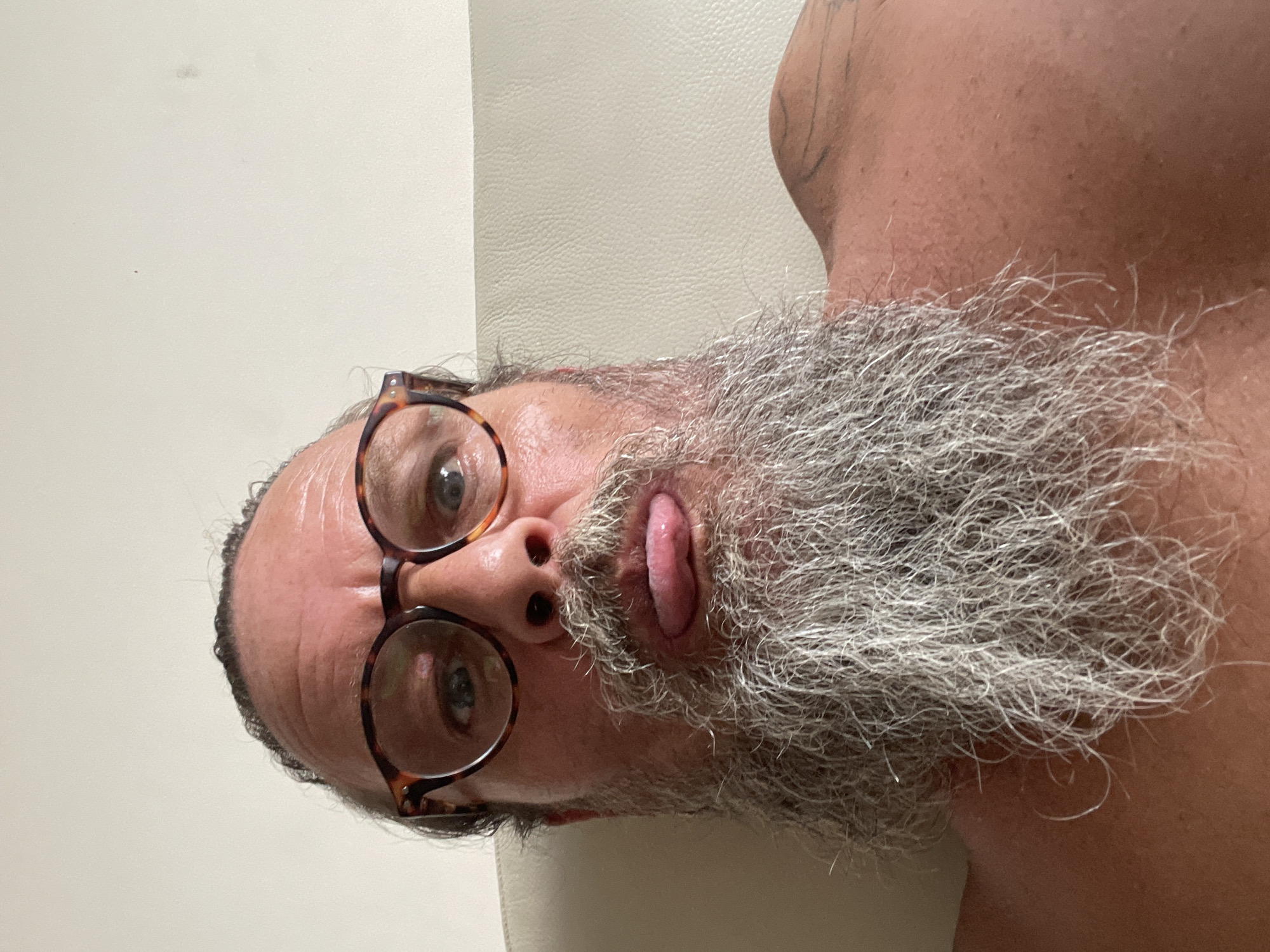 A man with a full grey beard, wearing tortoise-shell glasses, sticks his tongue out at the camera. His hair is pulled back. He is shirtless, and a tattoo is visible on his right shoulder. The background appears to be a plain, light-colored wall and a textured, light-colored surface that could be upholstery. The image appears to have been taken indoors.