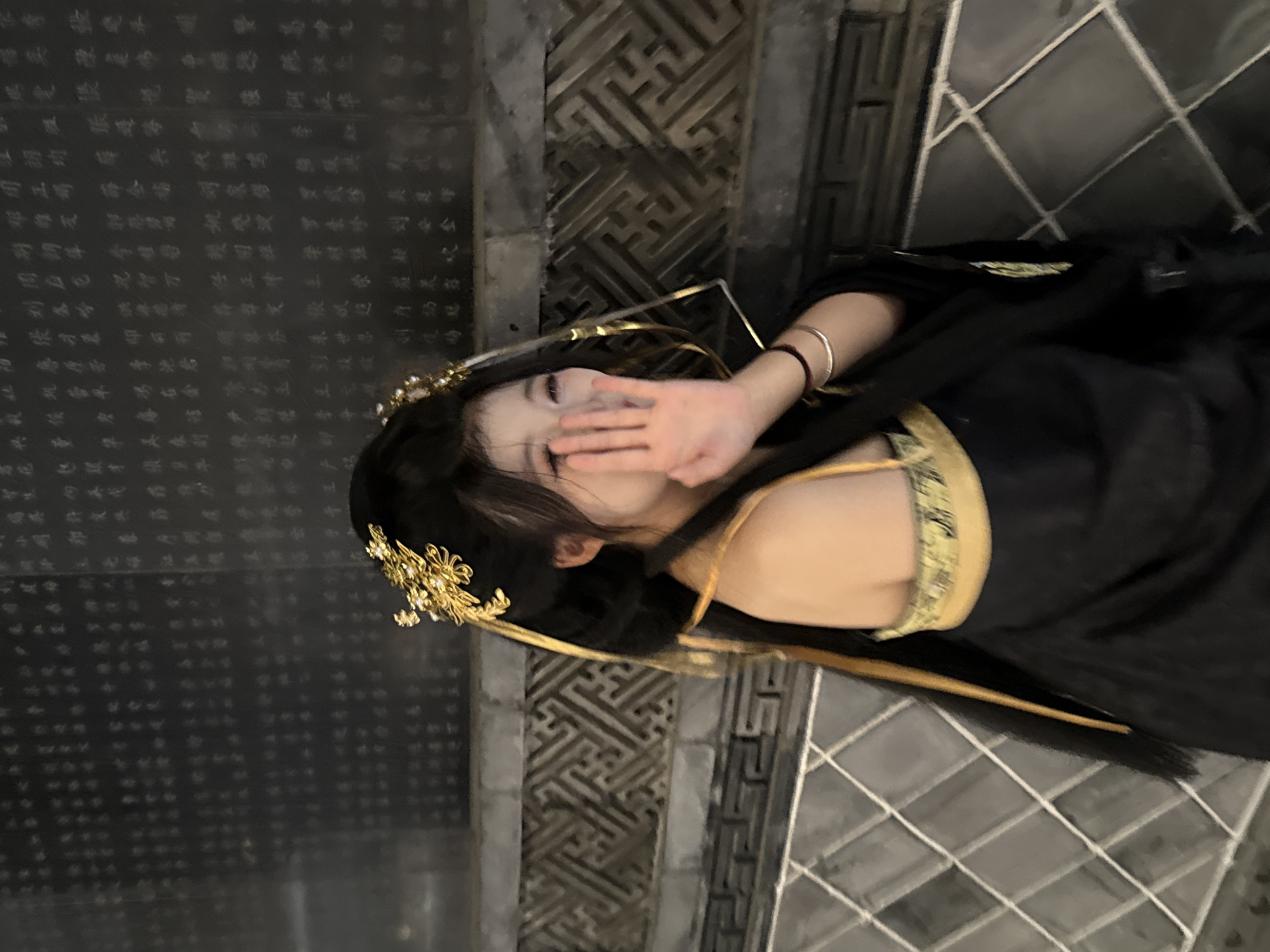 A woman in a black and gold dress with ornate gold hair pieces is lying on what appears to be a stone surface. She is covering her face with her hand. The background consists of patterned stone or tile surfaces. The setting appears to be an outdoor or semi-outdoor area with architectural elements that suggest a location in Yuzhong District, China. The black wall in the background to the left looks to be inscribed with Chinese characters.