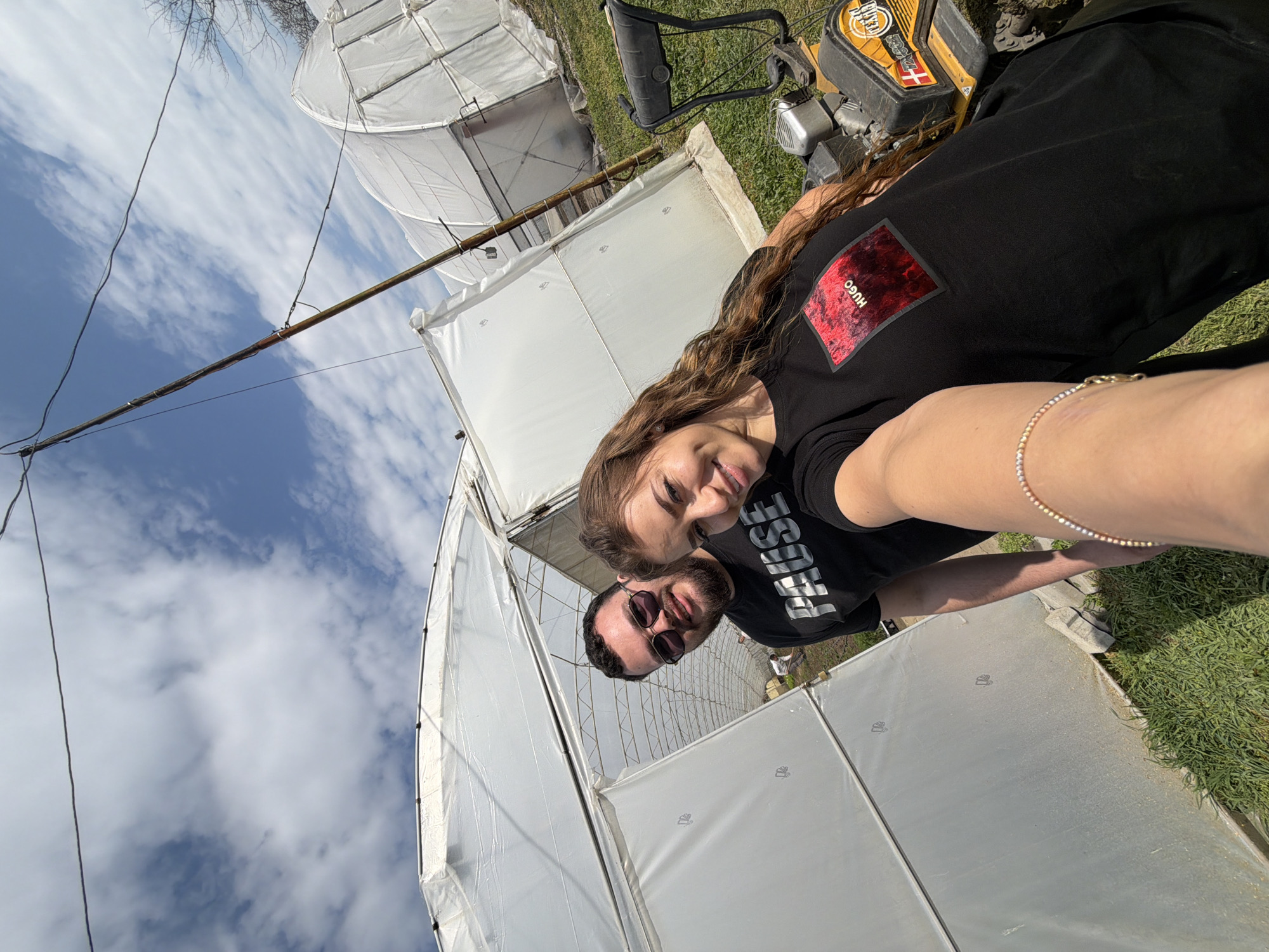 The image depicts two individuals, a male and a female, in an agricultural setting in Plovdiv, Bulgaria, characterized by multiple hoop-style greenhouses. The female, positioned in the foreground and appearing to take the selfie, has long, wavy brown hair and wears a black t-shirt with a red "HUGO" graphic on the chest. A multi-colored beaded bracelet is visible on her right wrist, which is extended towards the camera. She smiles directly at the viewer. To her left and slightly behind, a male subject with a beard and sunglasses wears a black t-shirt displaying "PAUSE" in white text. He also smiles towards the camera.
In the background, several large greenhouses covered in translucent white plastic sheeting are visible under a partly cloudy blue sky. The entrance of the most prominent greenhouse, located directly behind the subjects, is open, revealing green plants and a blurred figure in a white top engaged in work inside. To the right of the female, a yellow and black piece of agricultural machinery, possibly a tiller, with "TEXAS" branding and a Danish flag logo, is partially visible on the grassy ground. Overhead, thin black wires traverse the sky, and a tall, slender pole, likely a support structure, rises on the right side of the frame. The immediate ground is covered in green grass and some exposed soil.
