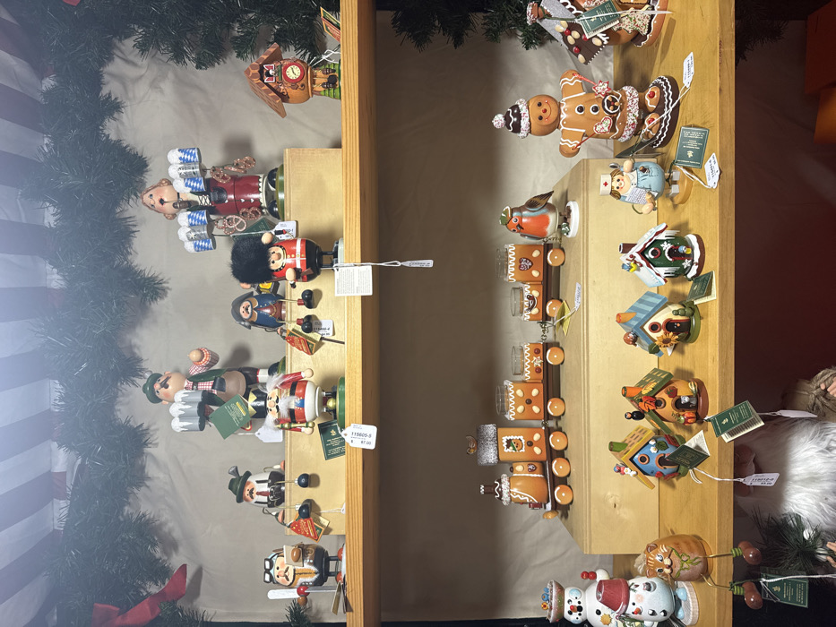 A shelf displays Christmas ornaments. On the left, are German nutcrackers. On the right, a gingerbread train, decorative birdhouses, and snowmen. The shelf is wooden and partially framed by a green garland. Tags hang from the ornaments, indicating their prices. Some items depict people, like the gingerbread man, nutcracker soldiers, and a nurse. Other items are nature-themed, like the birdhouses, cat, and snowmen.