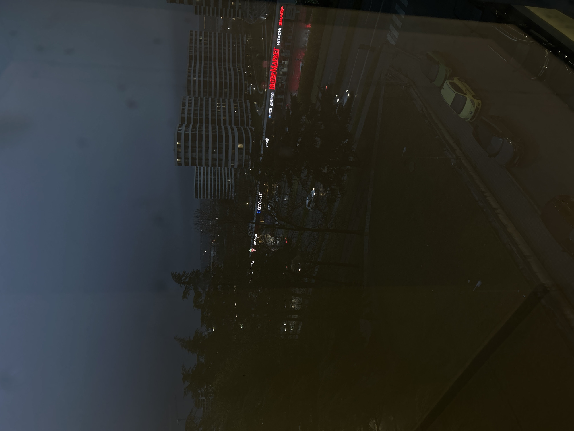 The image appears to be a night view from a window, possibly reflecting elements of the interior. Visible through the window is a street in Varna, Bulgaria. A large building complex, likely a residential or commercial development, stands prominently. A bright sign reading "INTER MARKET" is visible, along with the names of brands "HITACHI" and "SHARP". Below the building, trees are present. On the street, several cars are parked or moving.