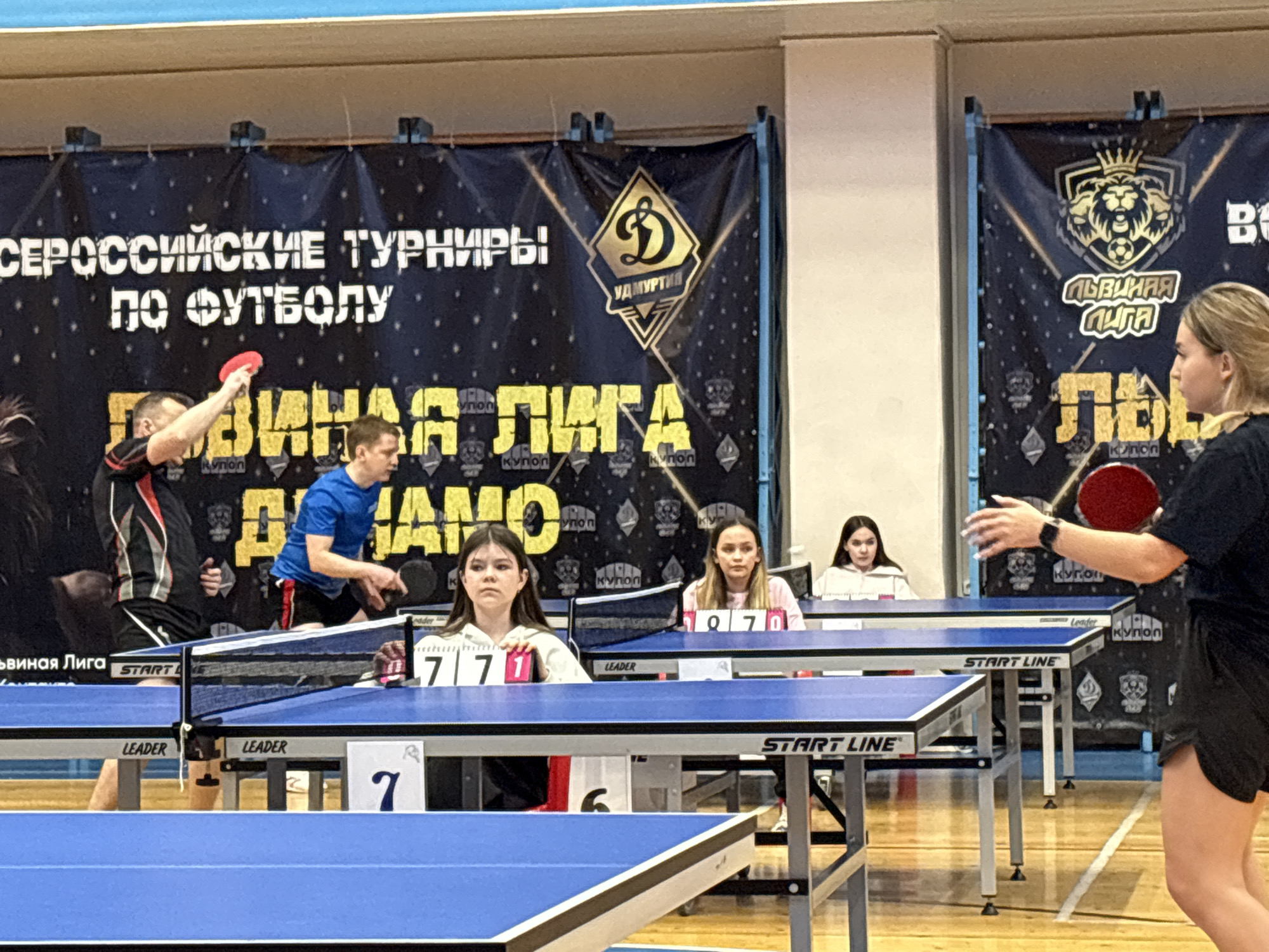 The image depicts a table tennis tournament, likely located in Izhevsk, Russia, based on the banners in the background. The banners display the text "СЕРОССИЙСКИЕ ТУРНИРЫ ПО ФУТБОЛУ" (All-Russian Football Tournaments) and "ЛЬВИНАЯ ЛИГА" (Lion League) along with some logos. Four people are playing table tennis: a man in black and red is about to hit the ball with his paddle, and a woman in black is preparing to receive. A man in a blue shirt is playing on the table next to them. Additionally, there are three female referees sitting behind the tables. Each table has a score indicator displaying numbers. The hall has a wooden floor.