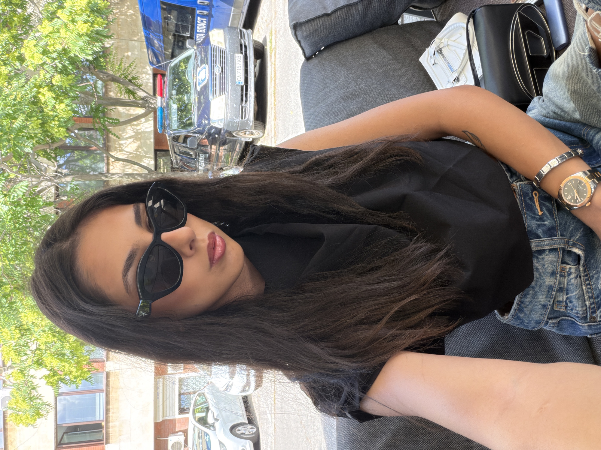 A young woman with long, dark hair and sunglasses is taking a selfie. She is wearing a black top and denim shorts. She is sitting on a gray couch outside, with a building and trees in the background. There are cars and a police van visible in the background. The police van has the license plate CA 7157 KB. The setting appears to be a street in the city of Dupnitsa, Bulgaria.