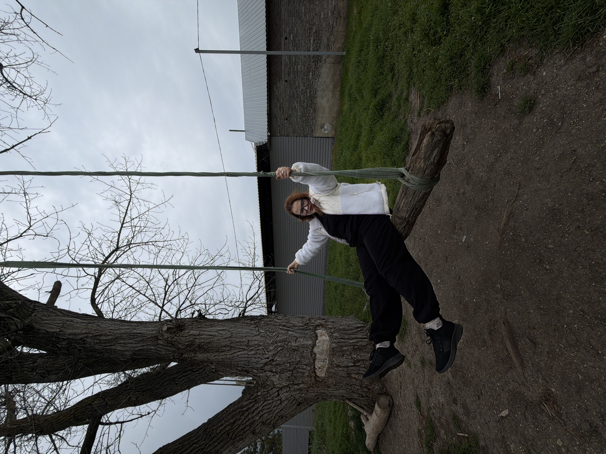 The image captures a full-length portrait of a young woman standing on a homemade swing in an outdoor, semi-rural setting in Port-Katon, Russia. The woman, with long reddish-brown hair and glasses, is smiling broadly at the camera. She is dressed in a white, possibly sherpa or quilted, jacket over a dark top, black pants, and black sneakers with white socks. On her visible right sock, the text "OUT SMILE" is discernable.
She is balancing on a rough, weathered wooden log that serves as the seat of the swing, holding onto thick, braided green ropes that extend upwards. The swing is suspended from a large, old tree with deeply furrowed bark and numerous bare branches, suggesting late winter or early spring. The sky overhead is overcast and a uniform light gray, indicating a cloudy daytime.
In the background, behind the woman and the tree, there is a simple, low-rise building with a corrugated metal roof and brick walls, set against patches of green grass. A utility pole is also visible near the building. The ground directly beneath the swing and around the tree is a mix of dirt and sparsely growing grass. The overall atmosphere appears casual and joyful, with the woman's smile being the focal point of emotion.