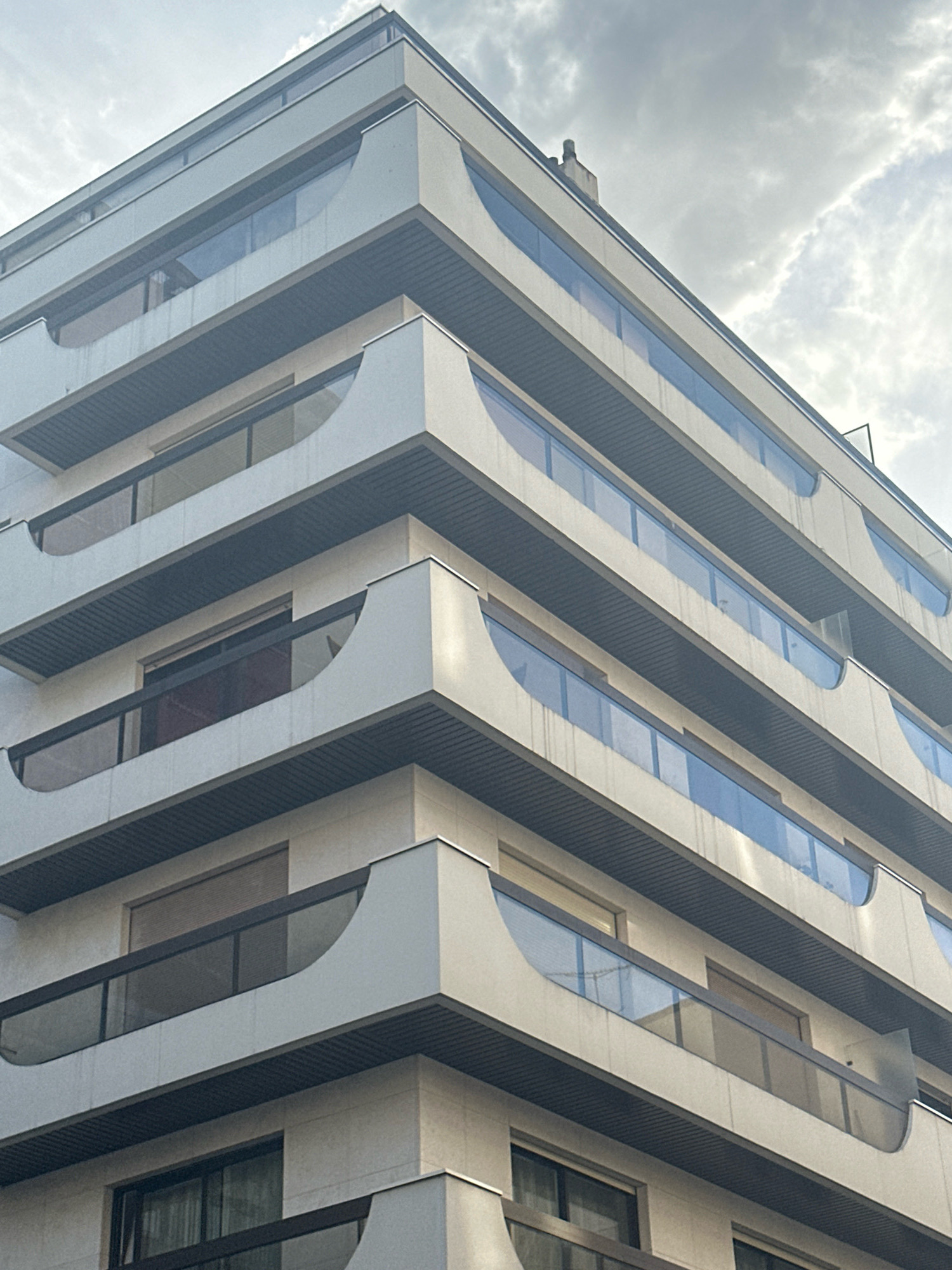 The image shows a close-up, low-angle view of the exterior of a modern apartment building. The building has multiple stories, each featuring a balcony with a curved glass railing and a textured, dark gray ceiling. The facade of the building is made of light-colored stone or concrete, with horizontal bands of darker material separating the floors. The balconies have a distinctive, rounded cutout at their outer edges. Above the building, a cloudy sky is visible. There are no people or vehicles in the frame. The building's architectural style suggests it is located in a contemporary urban setting, likely in a developed city. Paris, France, is known for its diverse architectural styles, and such a modern building could be found in various neighborhoods.